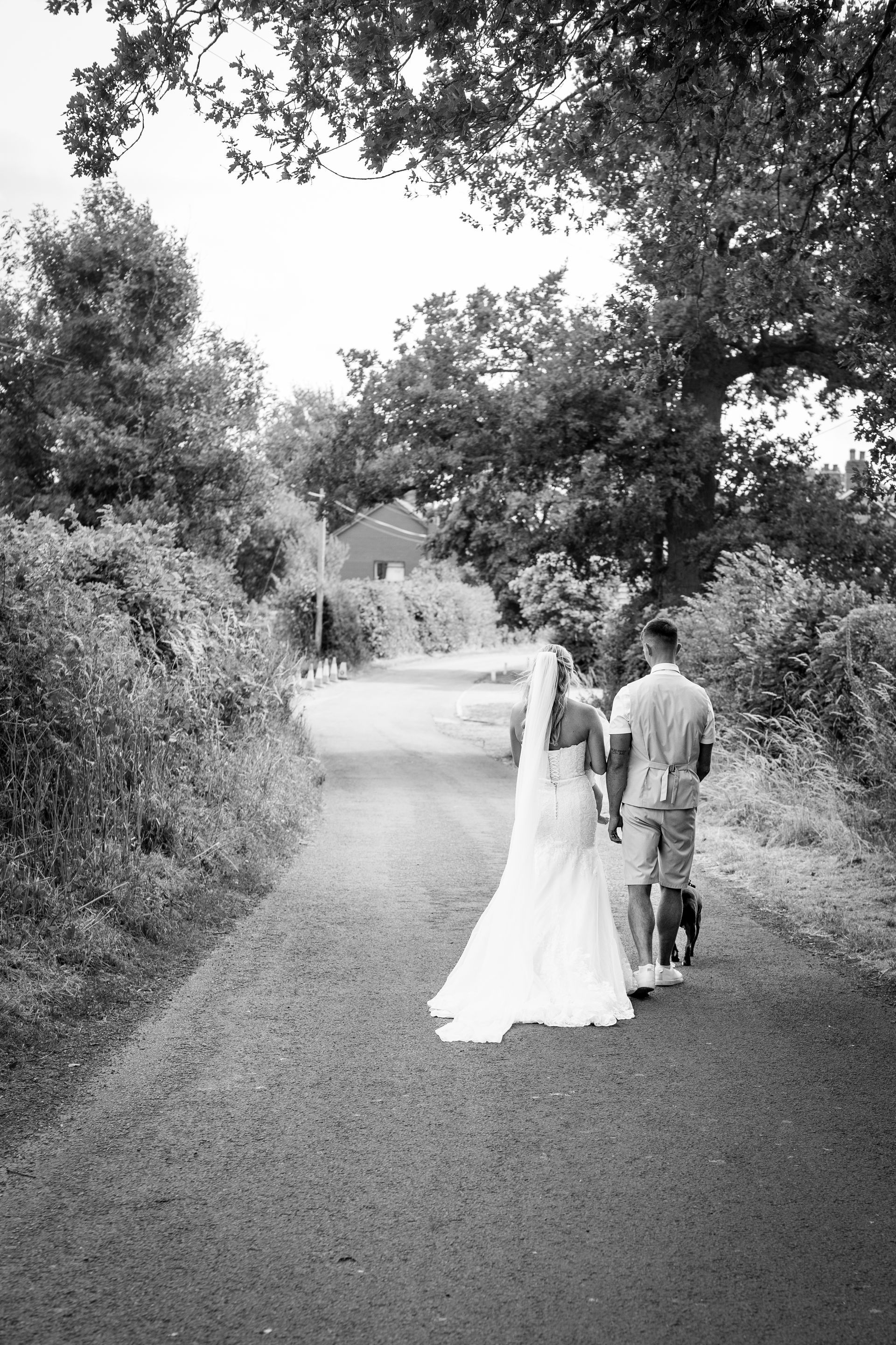 Bride and groom walk down a path in black and white. The bride wears a veil and dress; the groom wears shorts.