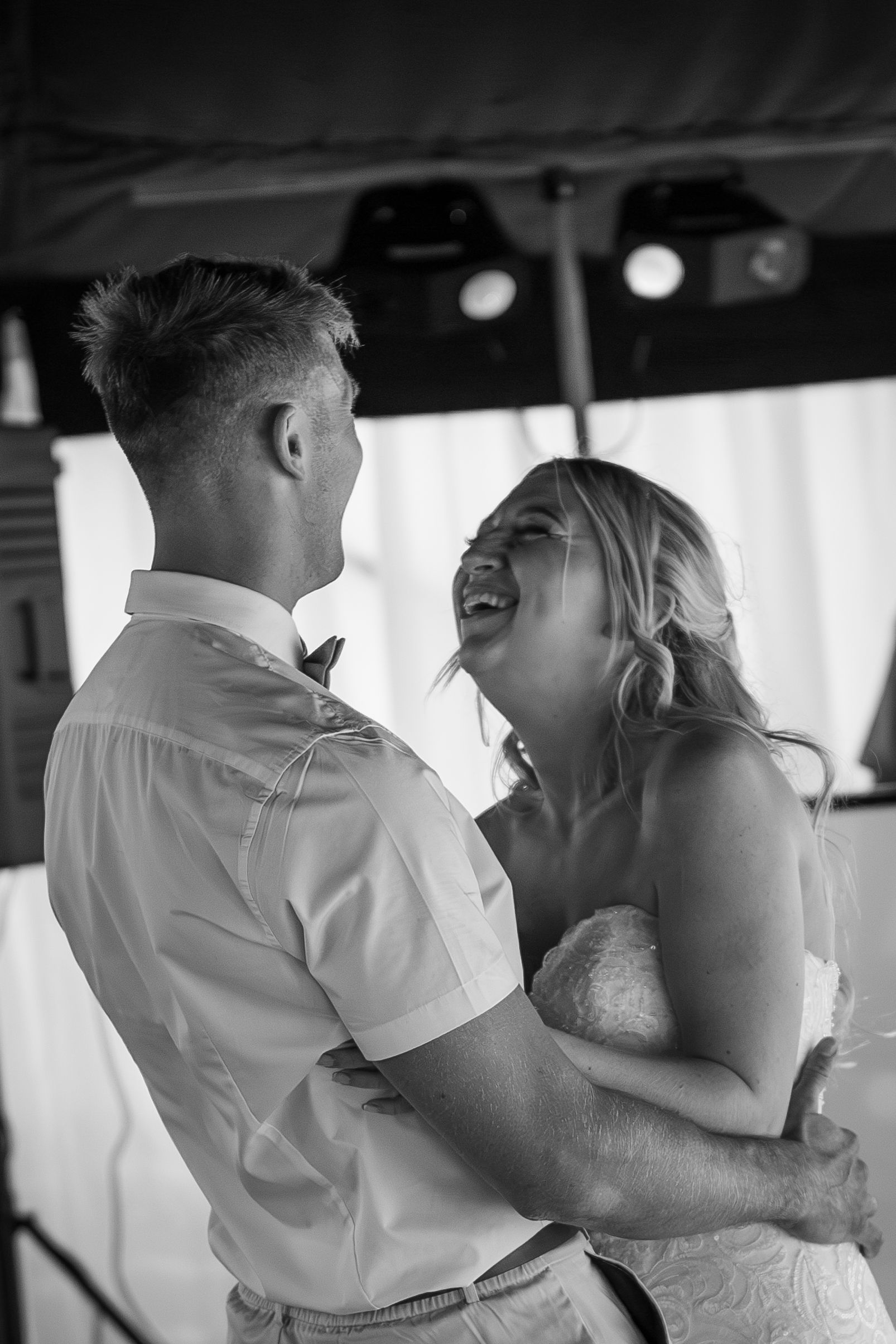 Bride and groom laugh while dancing at their wedding, with lights in the background.
