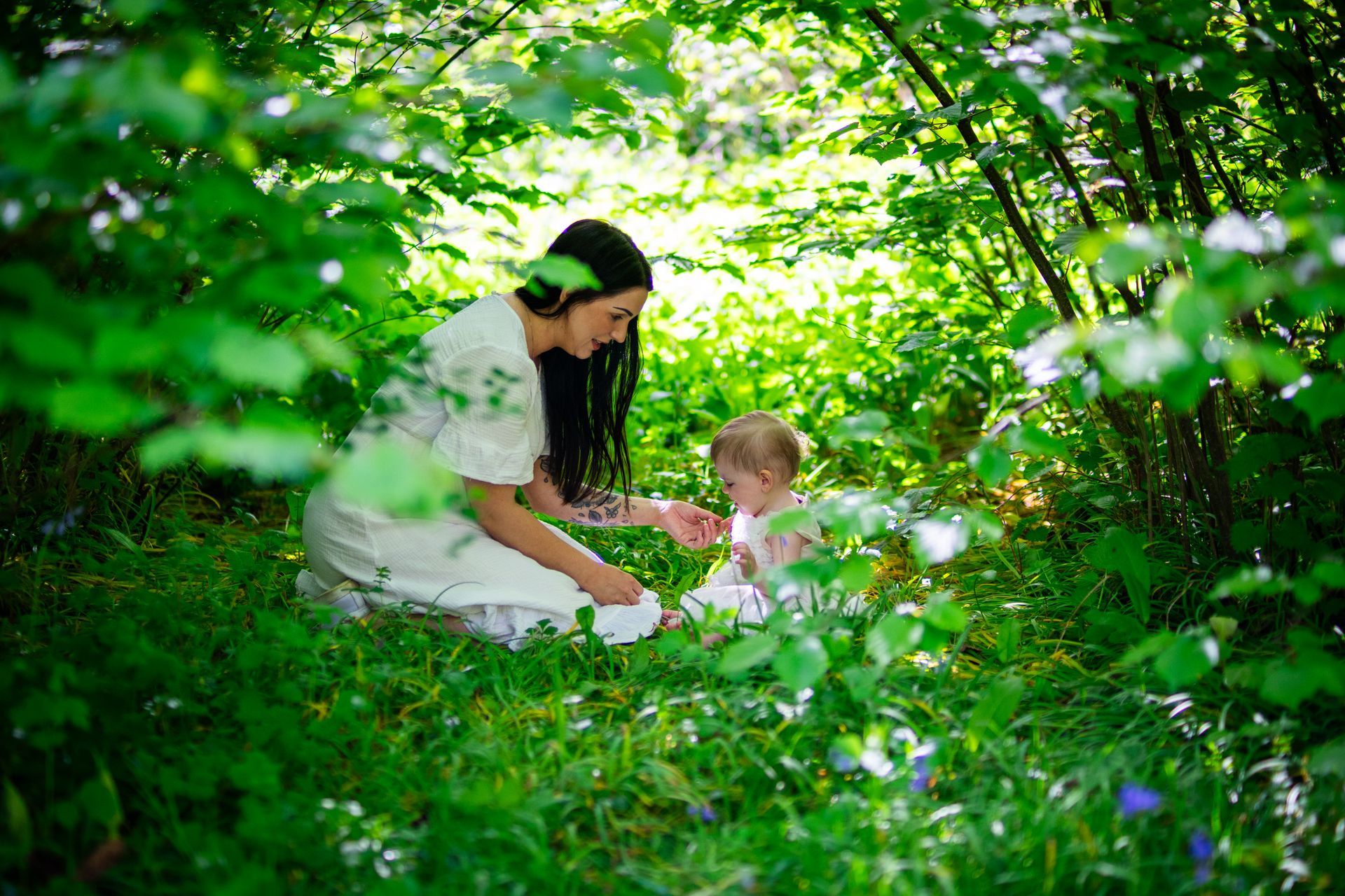 Woman and baby in white dresses sit in a lush green forest.