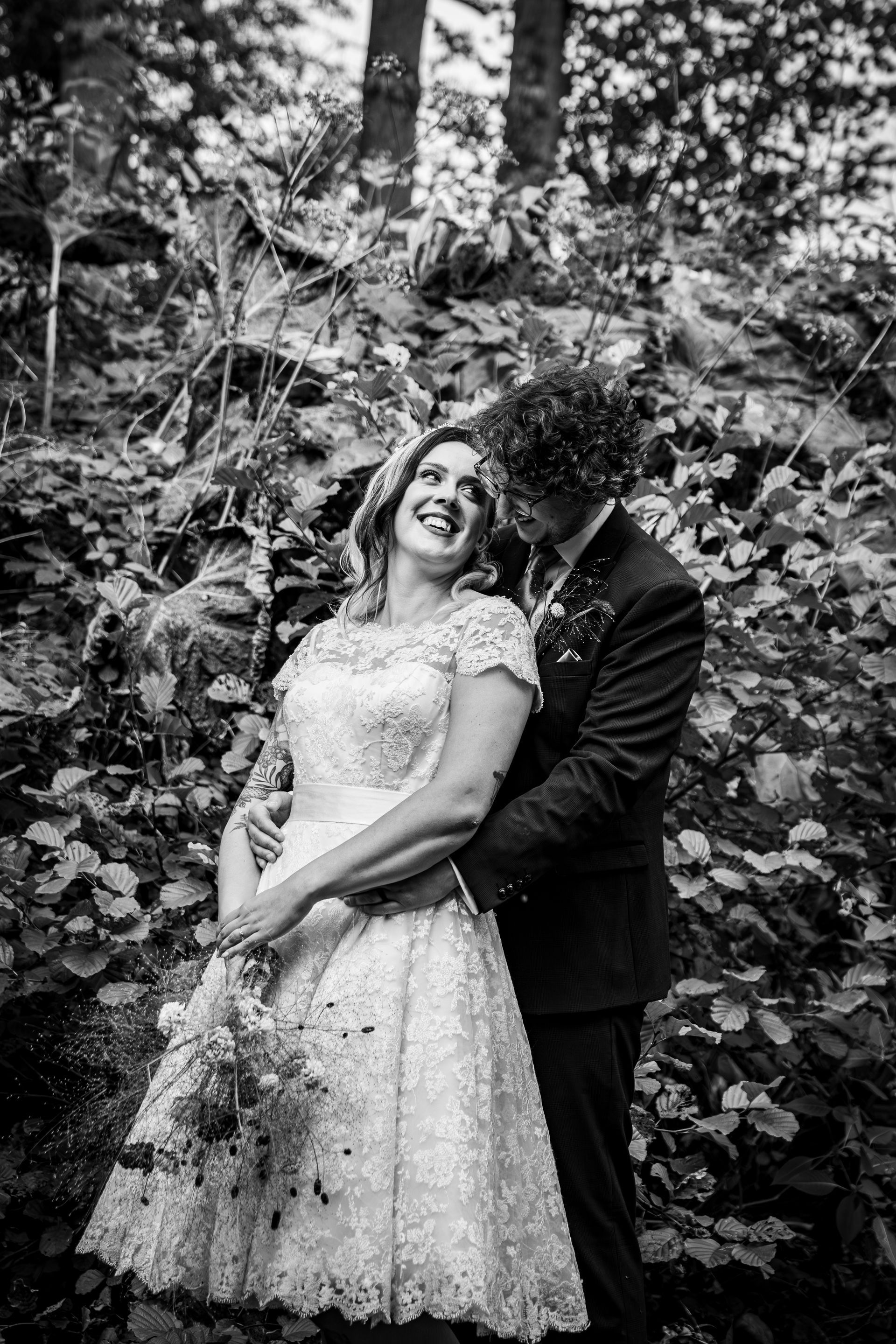 Bride in lace dress laughs as groom embraces her in a forest, black and white.