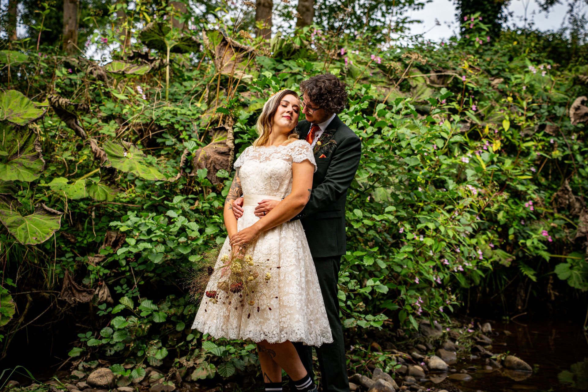 Couple embraces in a lush, green woodland setting; bride in a vintage-style dress, groom in a dark suit.