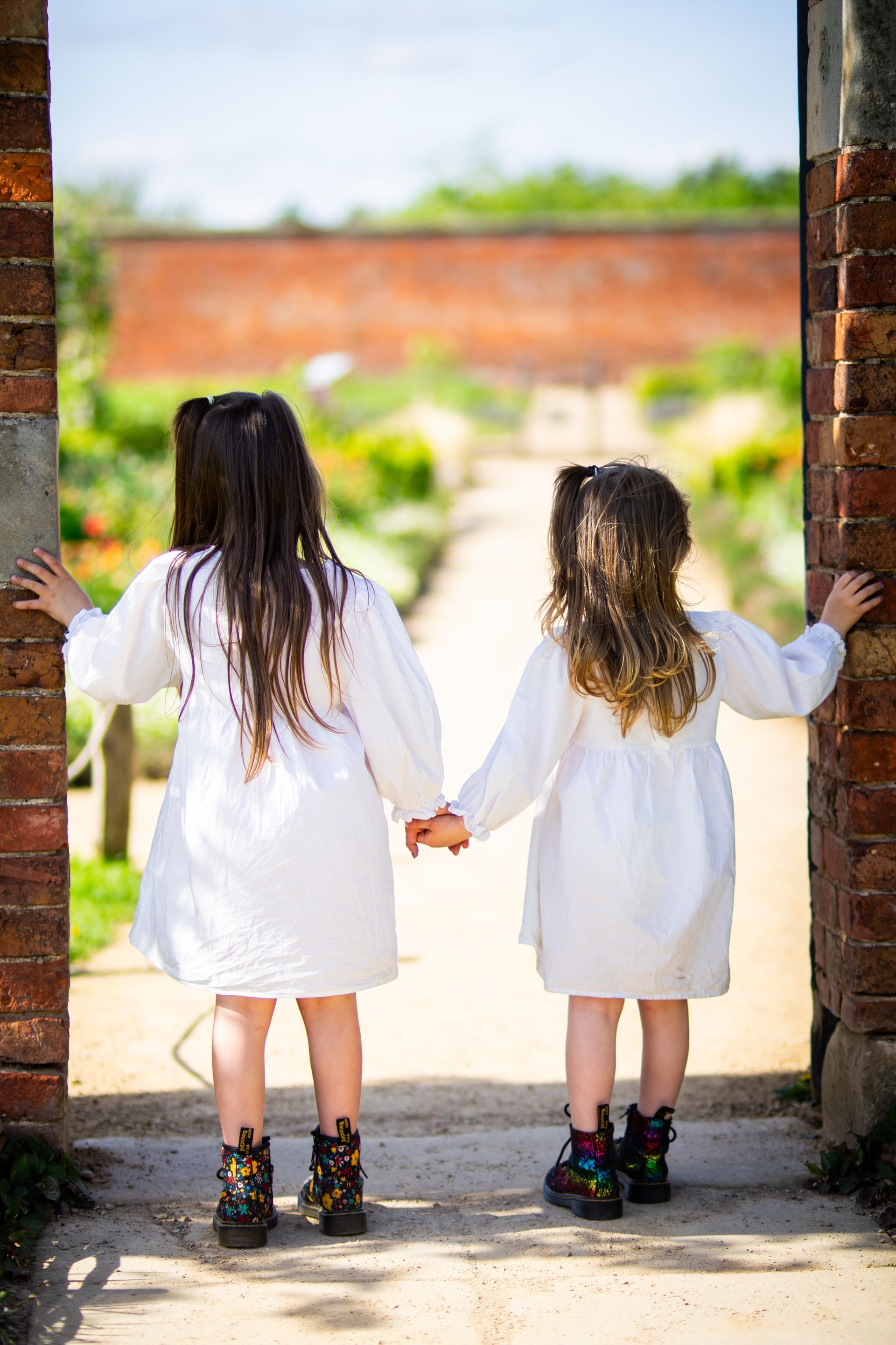 Two young girls in white dresses hold hands, gazing at a garden through a brick archway.