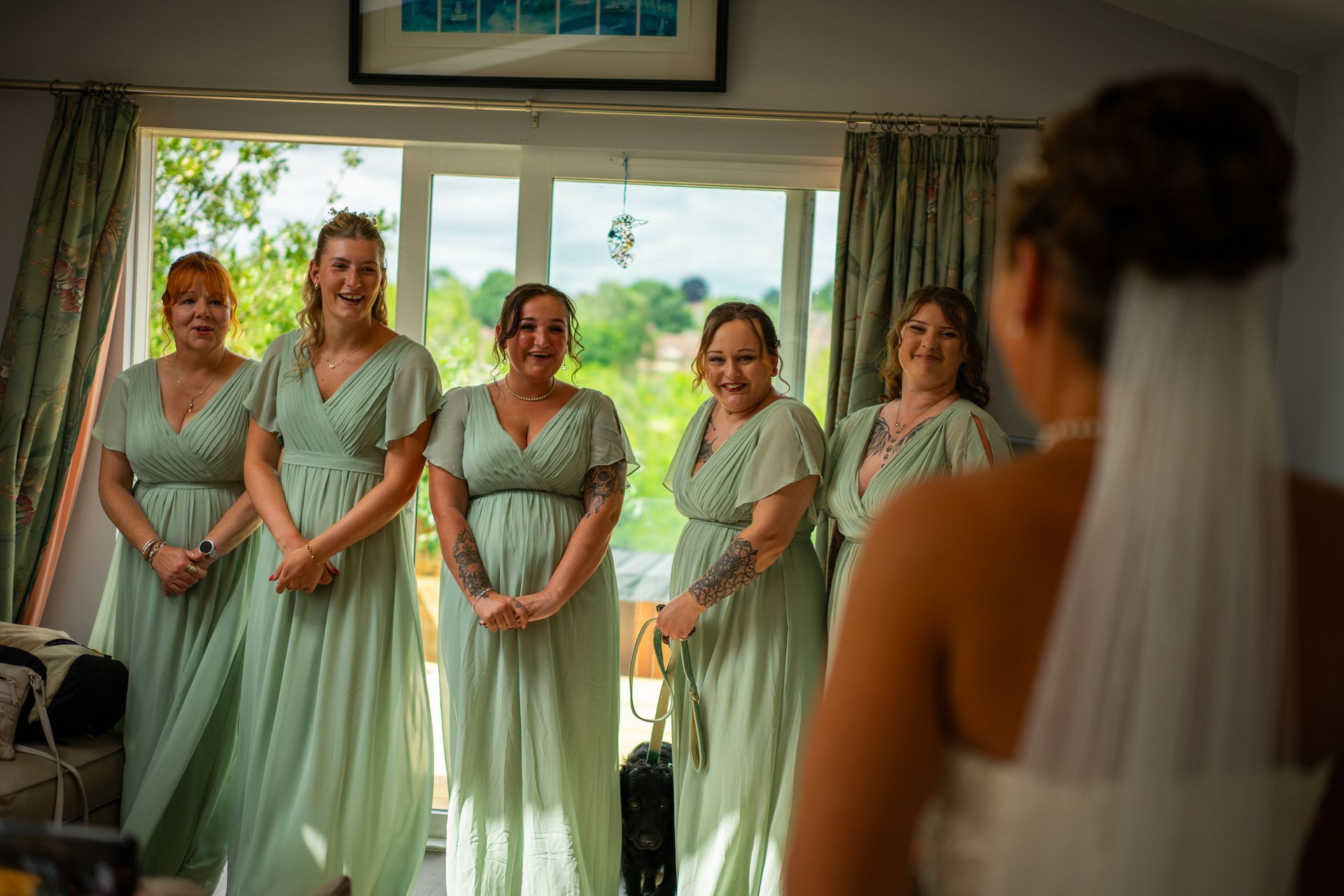 Bridesmaids in green dresses react to seeing the bride in her wedding dress. Sunlight streams through the window.