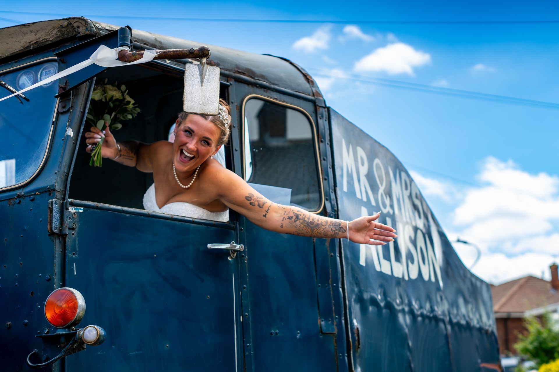 Bride in a blue truck, waving with flowers, arms out, smiling. 