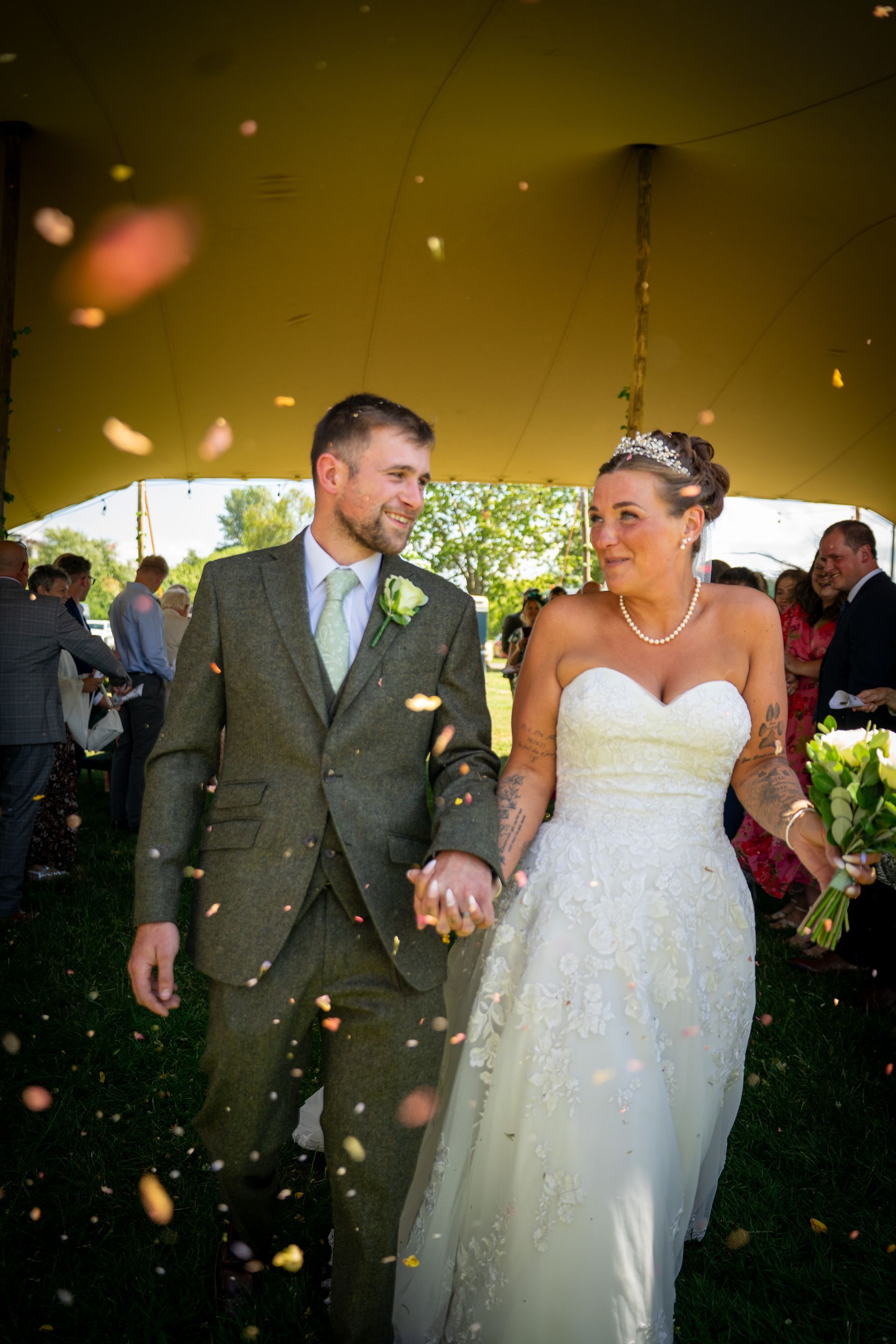 Newlyweds smiling, walking under a tent as flower petals fall; bride in white gown, groom in tweed suit.