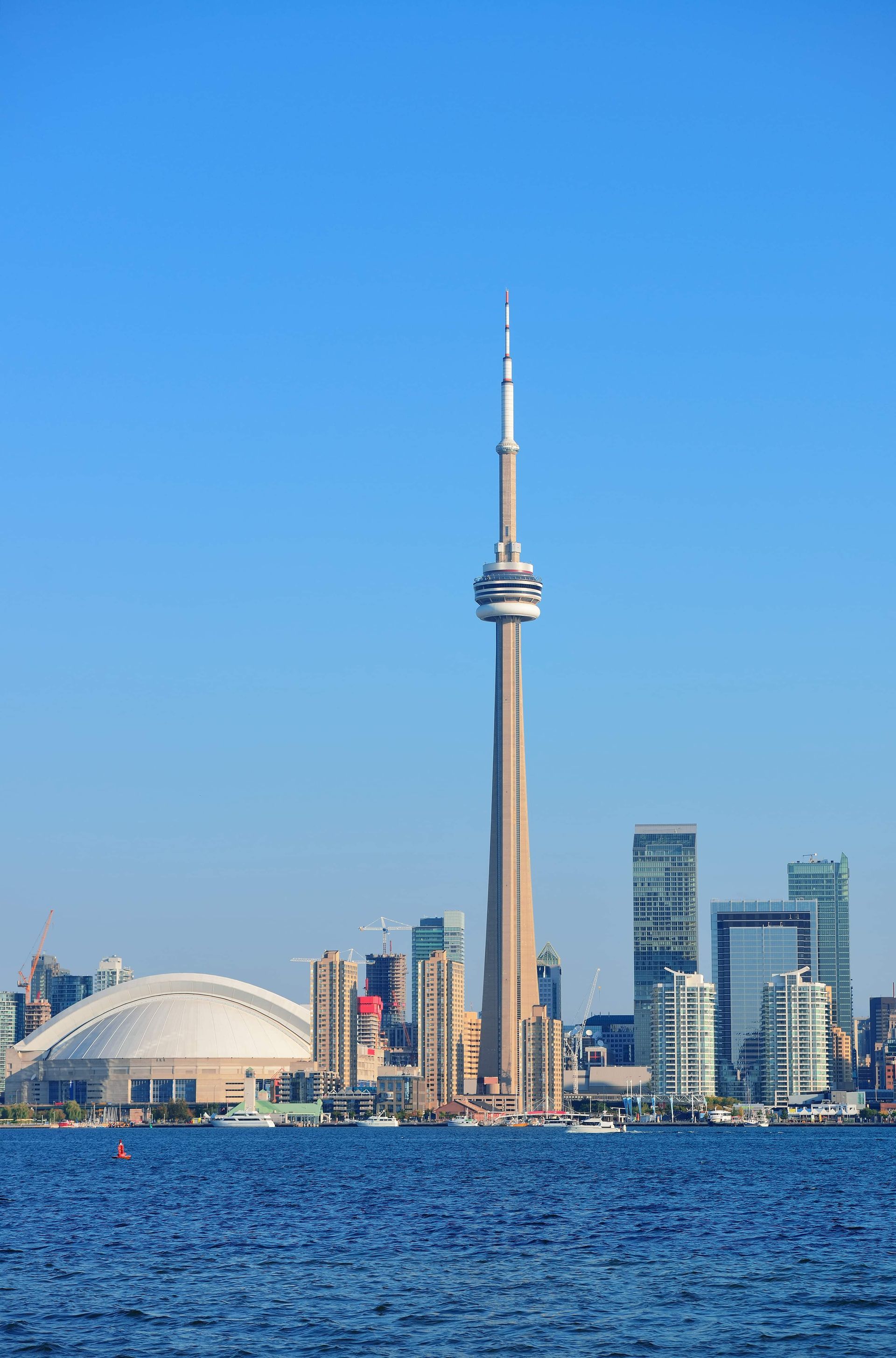 CN Tower and Toronto skyline, including a domed stadium, over a blue lake against a clear sky.