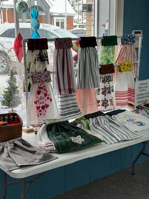 Display of decorative kitchen towels in a store window, arranged on a rack and table. Various colors and patterns are visible.