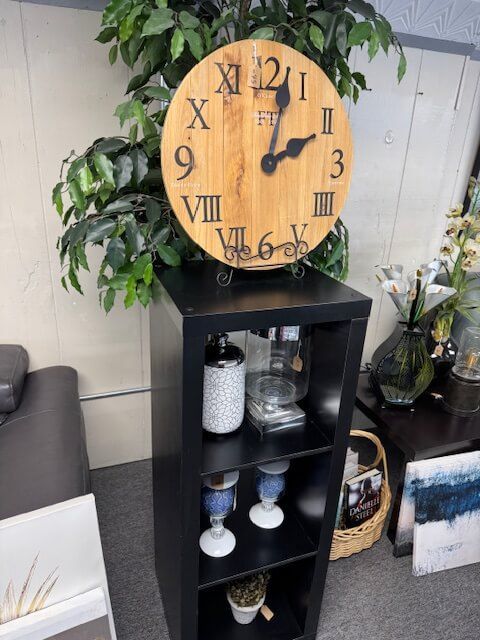 Clock with Roman numerals on a wooden face, sitting on a black shelf. Green plant in the background.