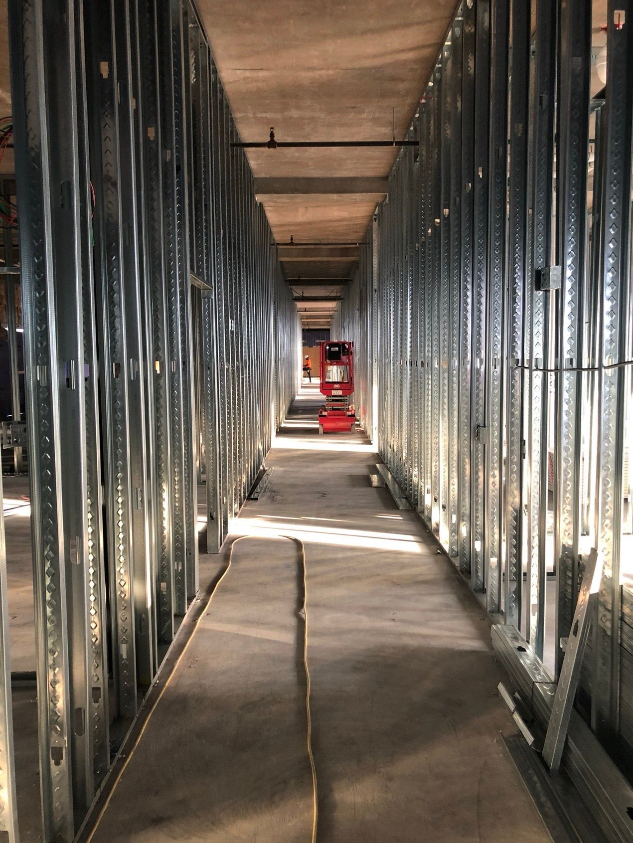 Metal studs form a long, narrow hallway in a construction site. A scissor lift sits in the distance.
