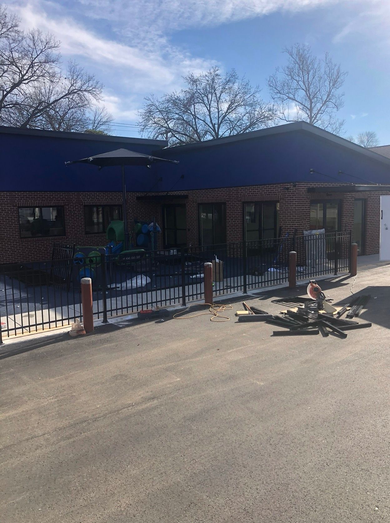 Building with blue roof and brick exterior, black fence, asphalt driveway, cloudy sky.