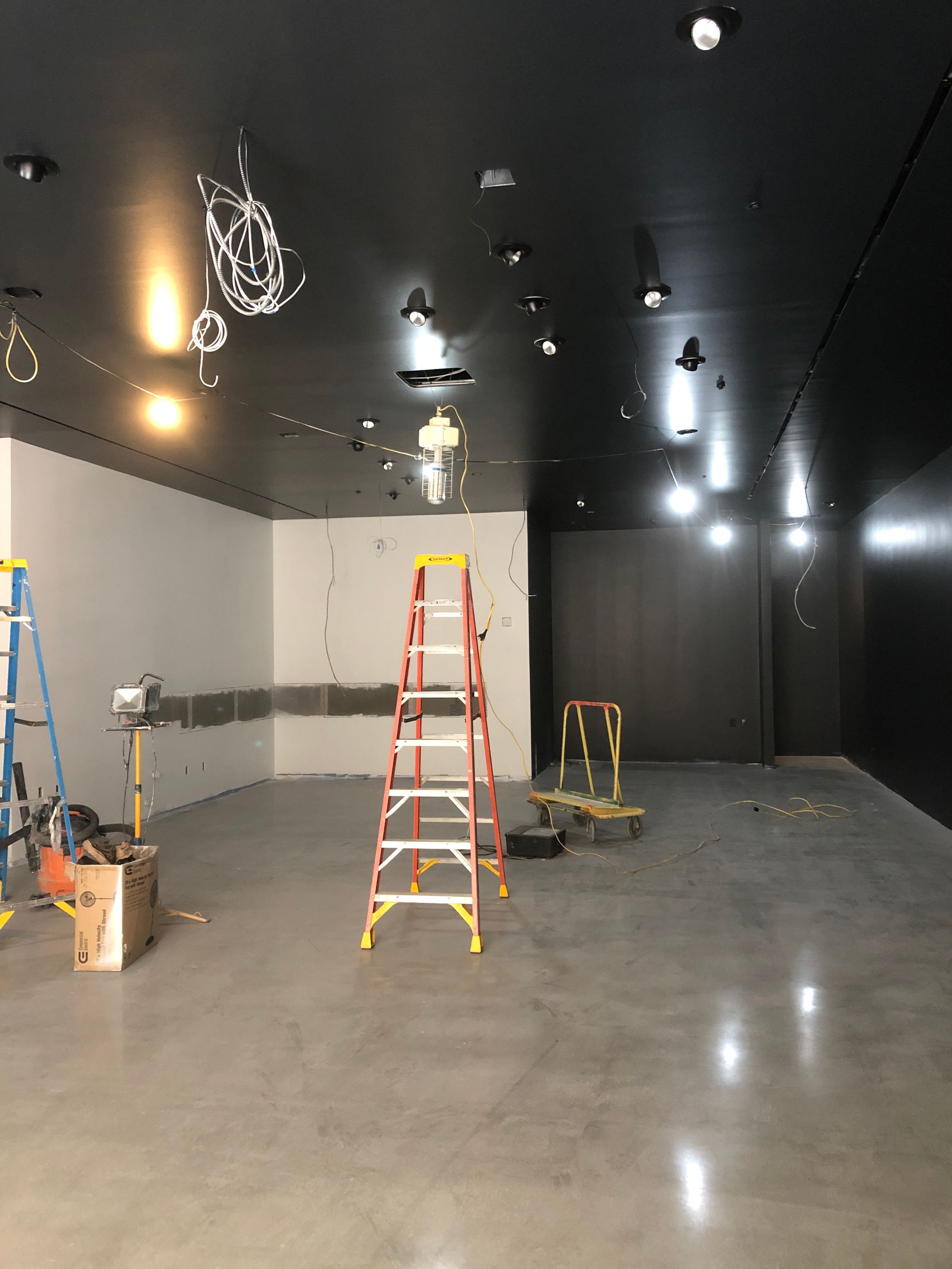 Interior of a room under construction with gray floor, black ceiling, white and dark gray walls, ladders, and exposed wiring.