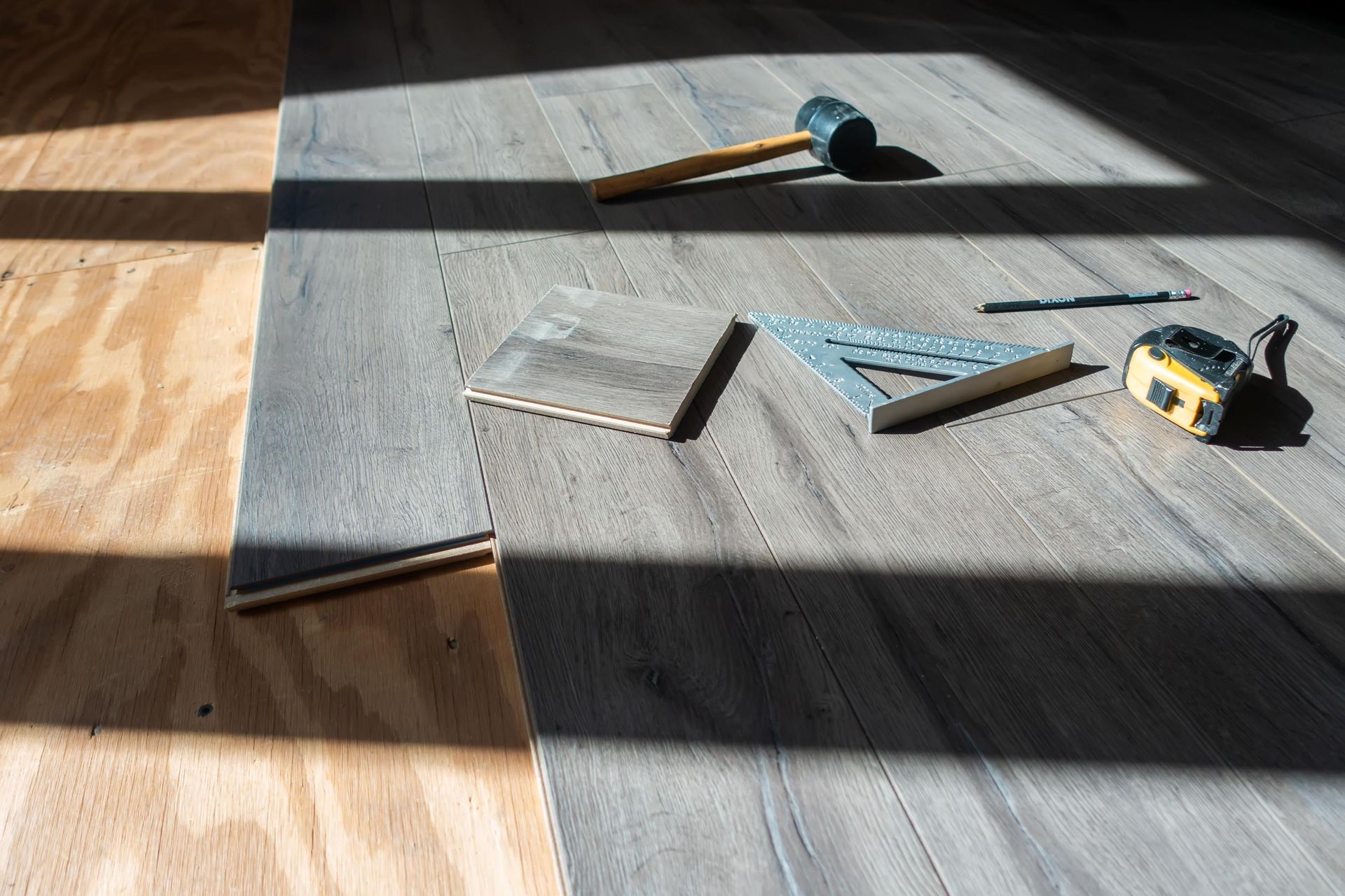 Tools and flooring materials on plywood: hammer, square, tape measure, pencil, partially laid gray wood planks.