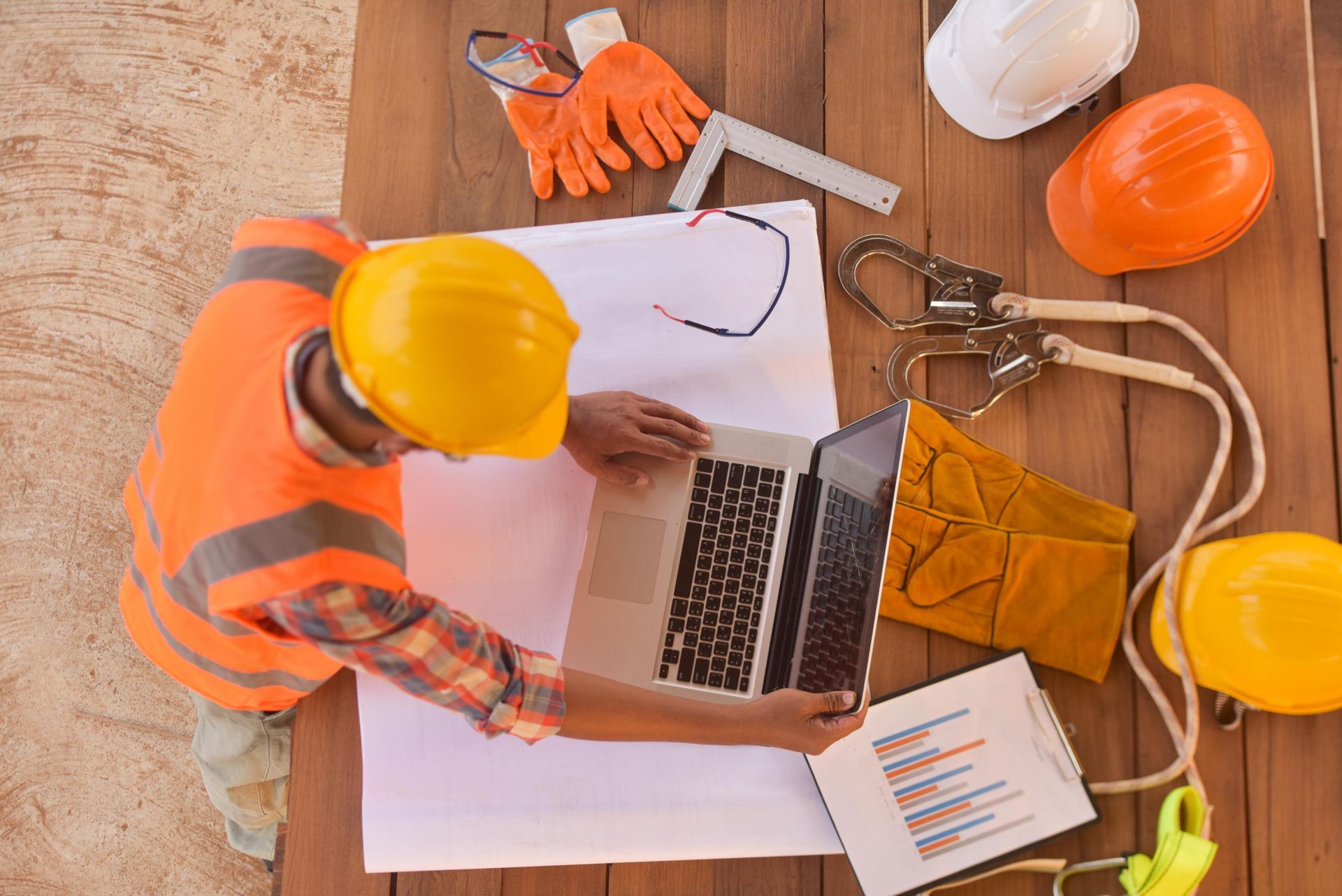 Person in safety vest and hard hat working on a laptop, surrounded by construction tools.