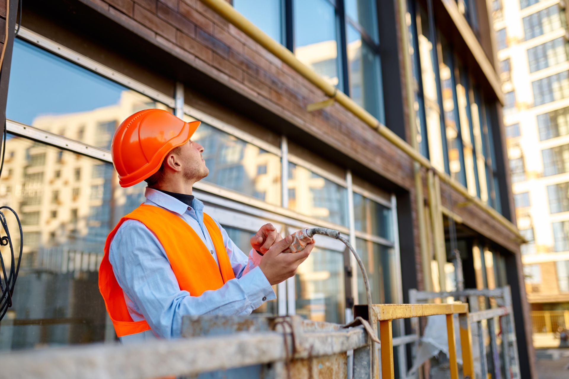Construction worker in orange safety gear inspects building exterior, holding a device.