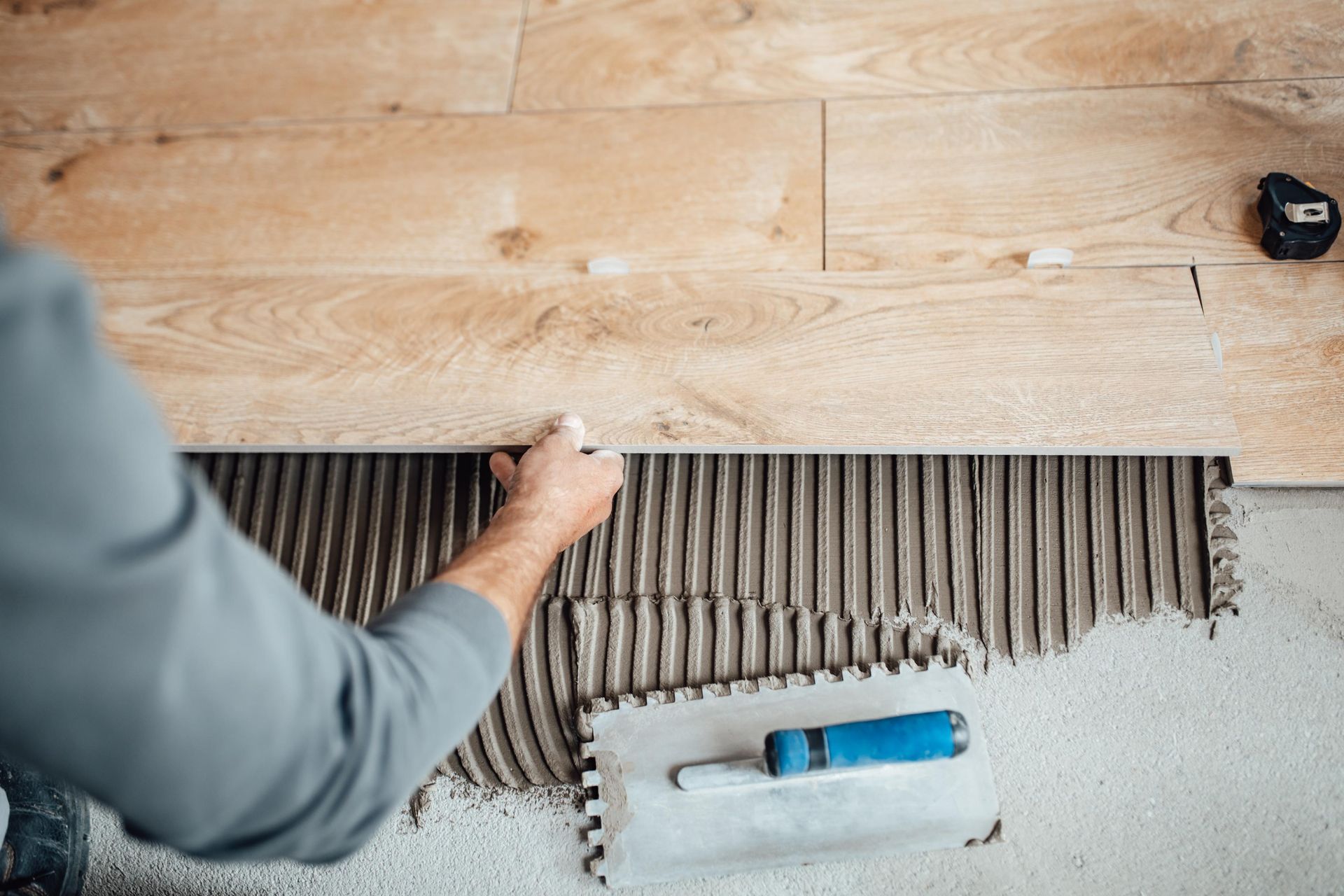 Person installing wood-look tile flooring, using a notched trowel and adhesive.