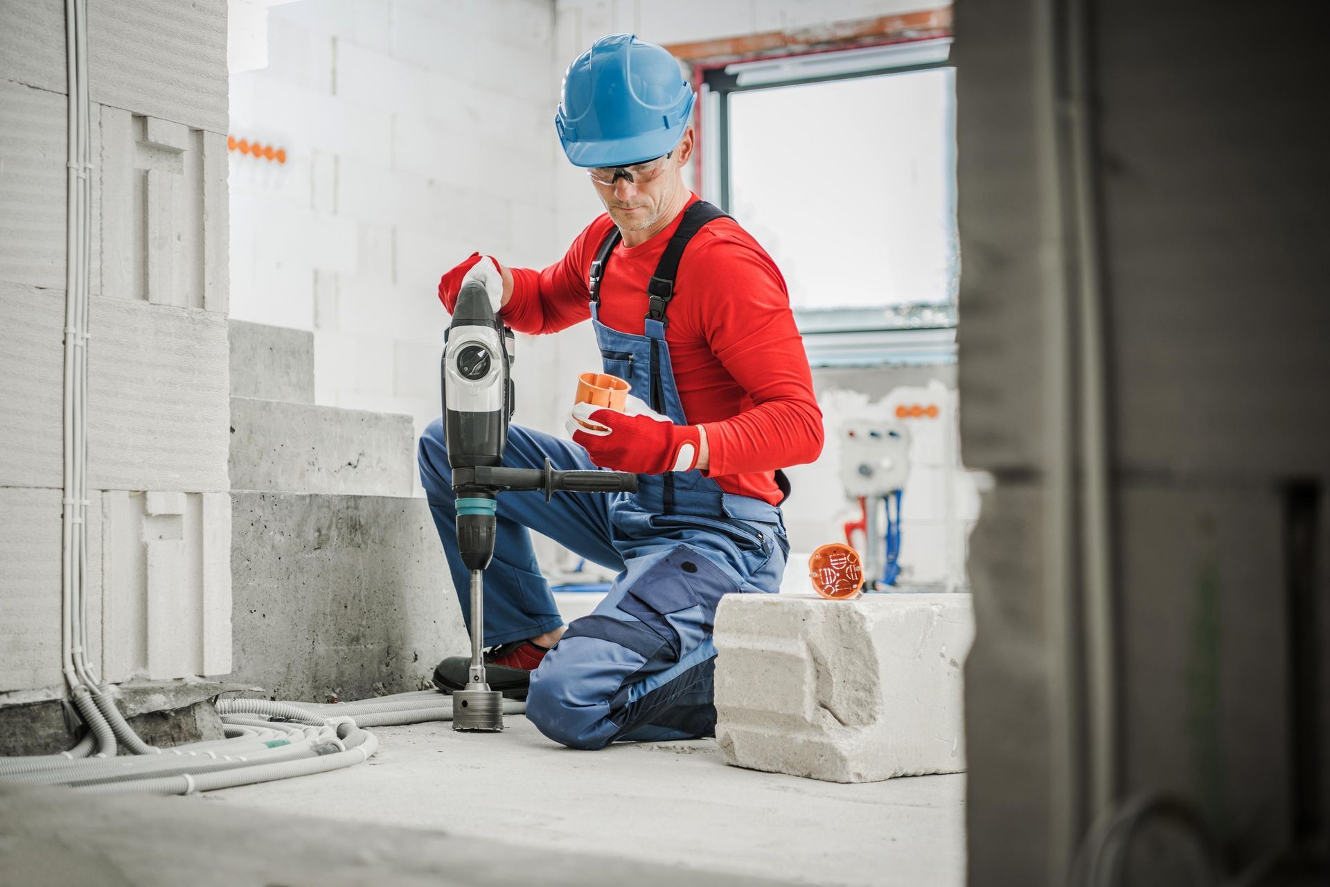 Construction worker drills into a concrete block while kneeling indoors, wearing blue overalls, red shirt, and hard hat.
