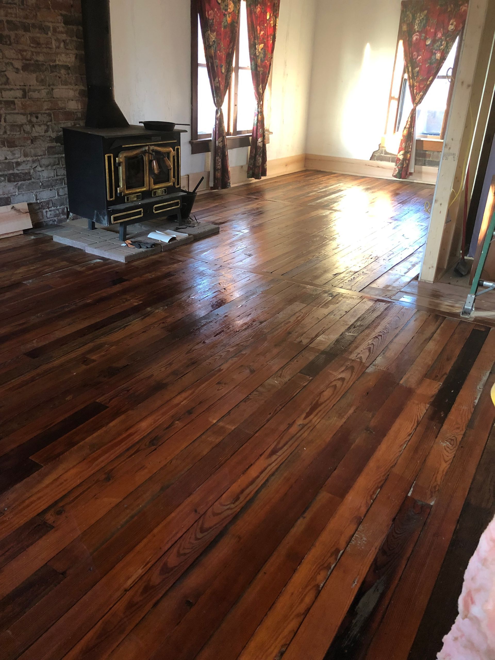 Refinished hardwood floor in a room with a wood-burning stove and curtains. Sunlight reflects off the wet surface.