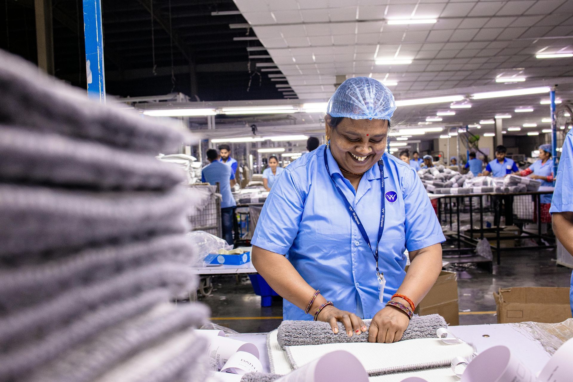 Woman in blue uniform smiles while working at a factory table, folding fabric.
