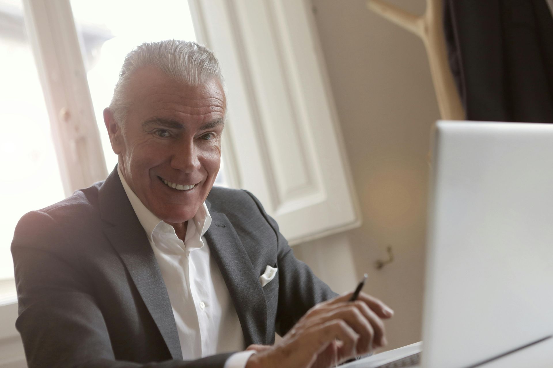 Smiling man in a suit working on a laptop near a bright window.