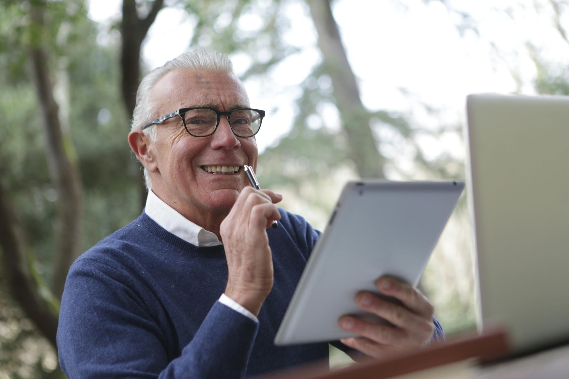 Smiling person with glasses holding a tablet and pen, sitting outdoors with a laptop.