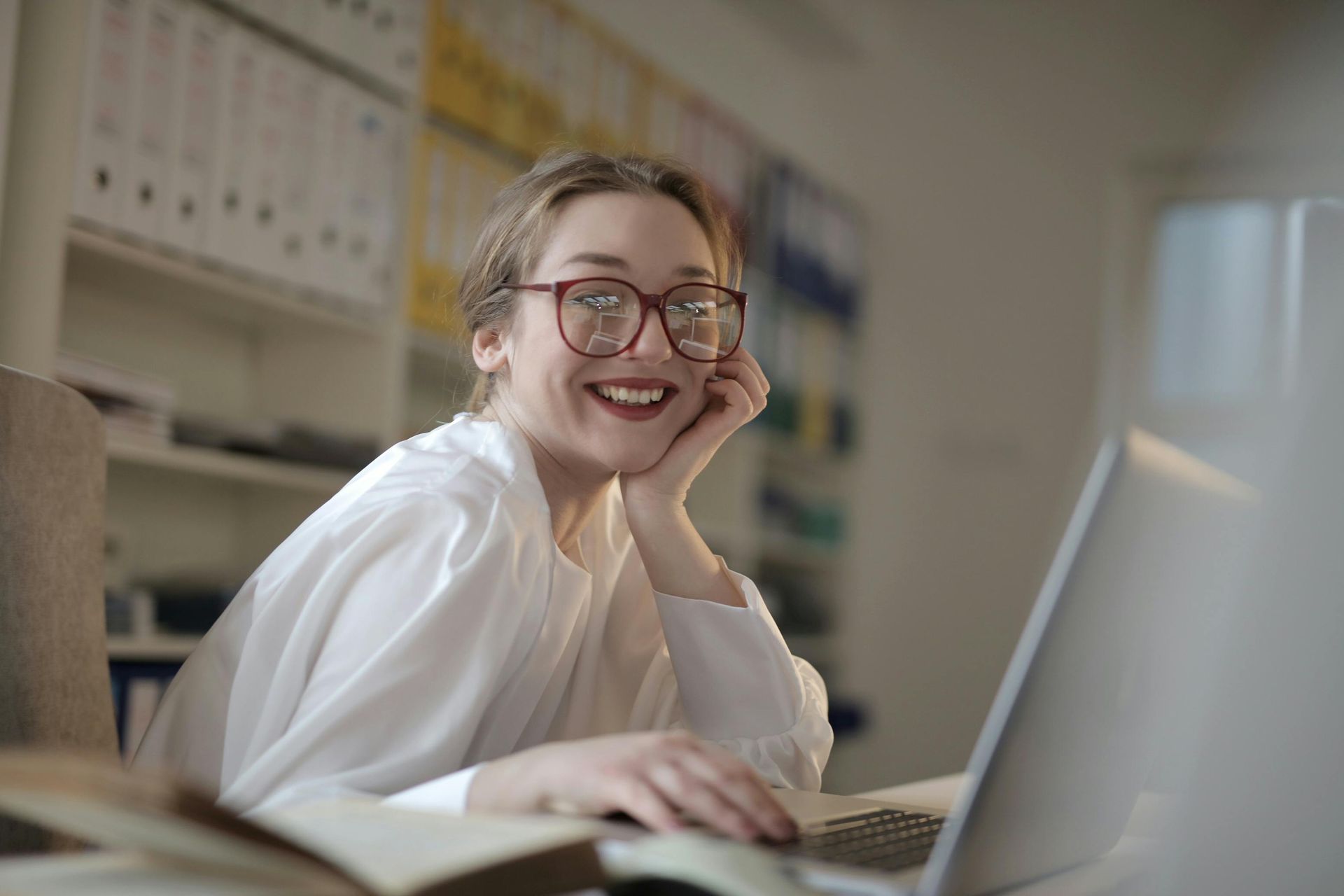 Woman with glasses smiles at the camera while working on a laptop. White shirt, office setting.