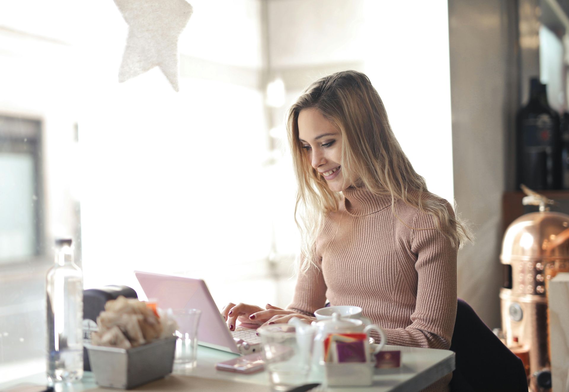 Woman in a pink sweater smiles, using a laptop at a cafe table near a window.