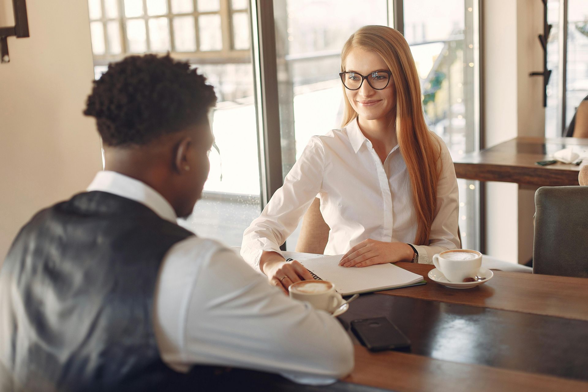 A woman in glasses interviews a person at a cafe table.