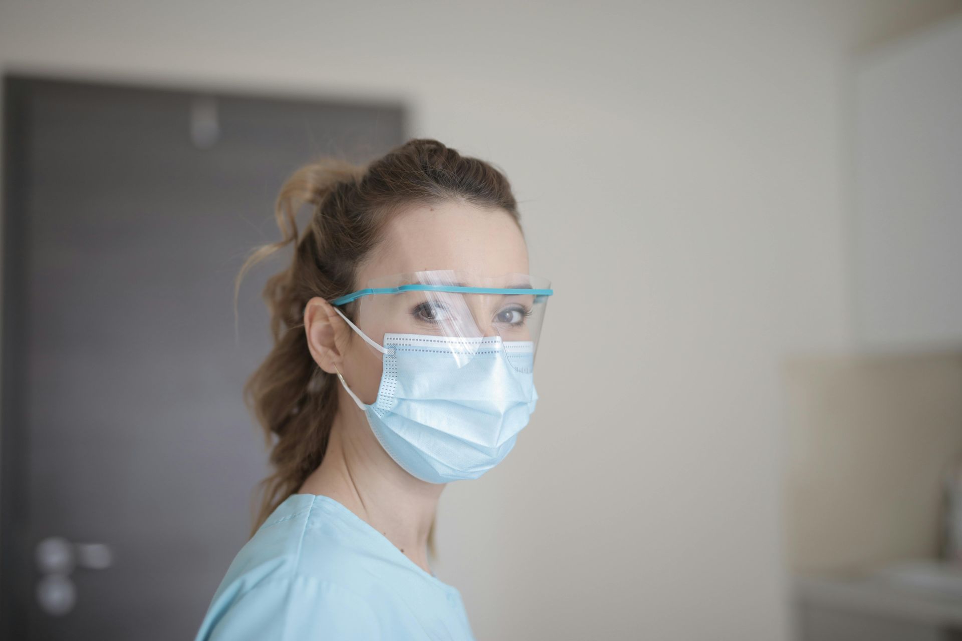 Woman wearing a surgical mask and face shield, looking toward the camera.