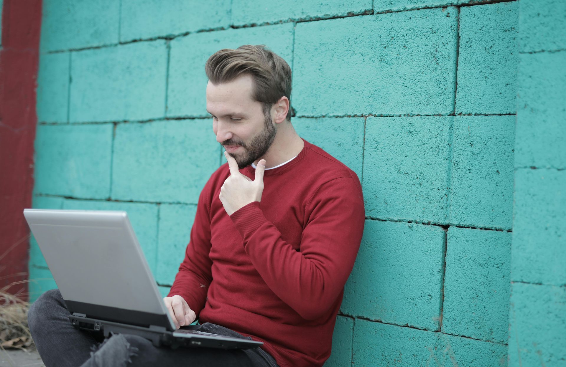 Man in red sweater using laptop, sitting against teal brick wall, thoughtful expression.