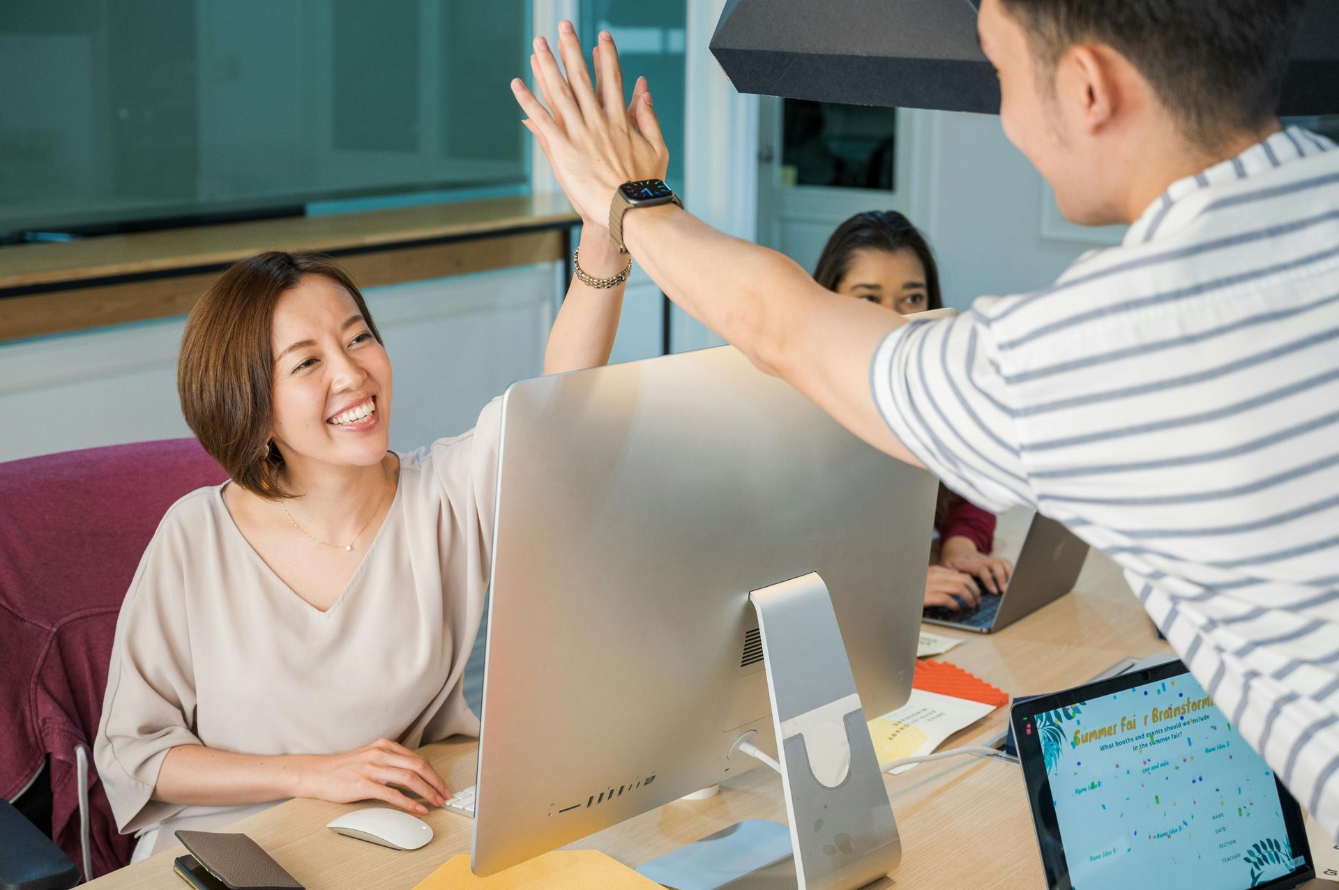Two people high-fiving over a computer. Smiling woman and man celebrate teamwork in an office setting.
