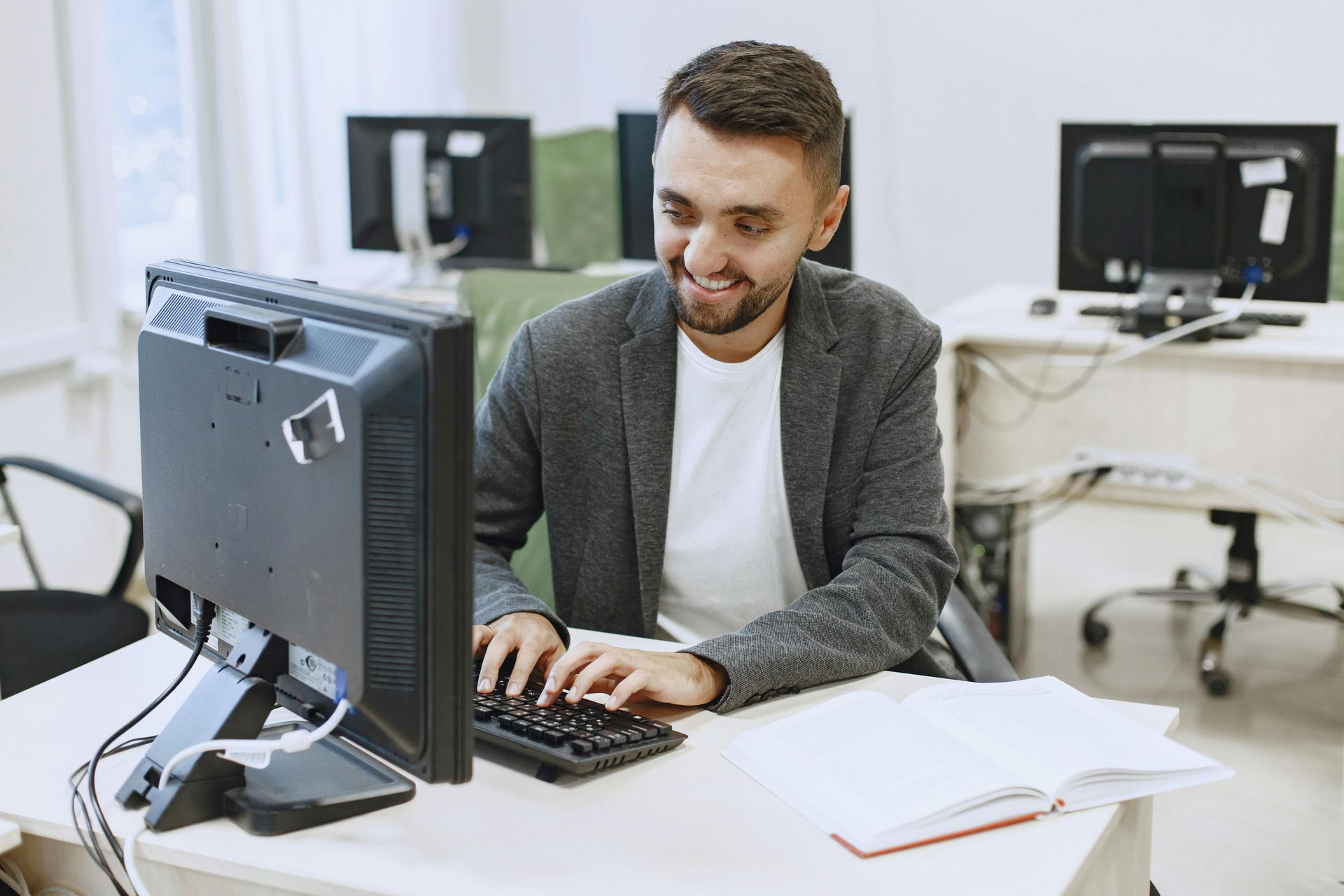Man smiling at computer keyboard in an office setting, with monitors and a notebook on the desk.