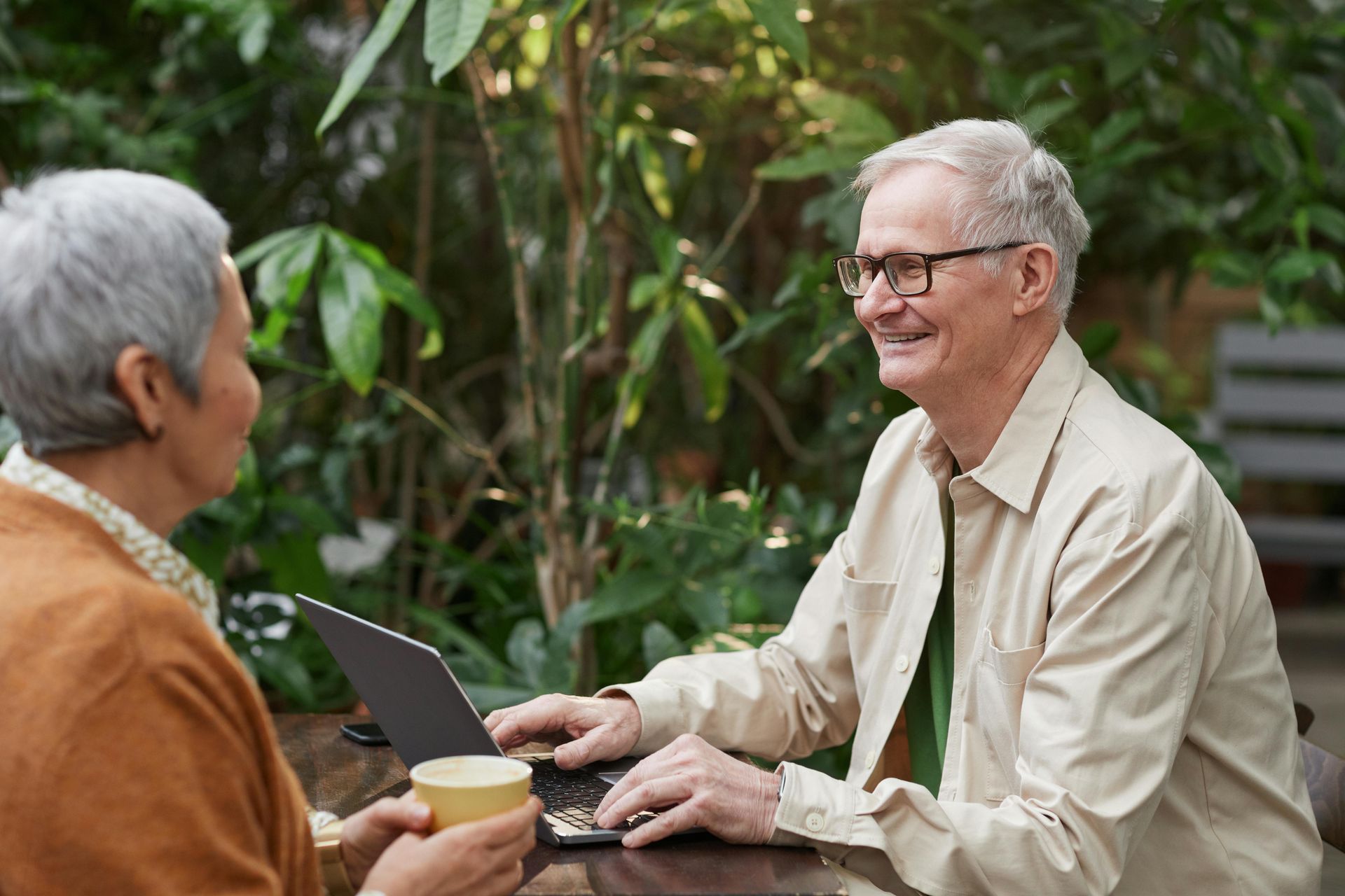 Two people at a table outdoors, one using a laptop, smiling. Green foliage in background.