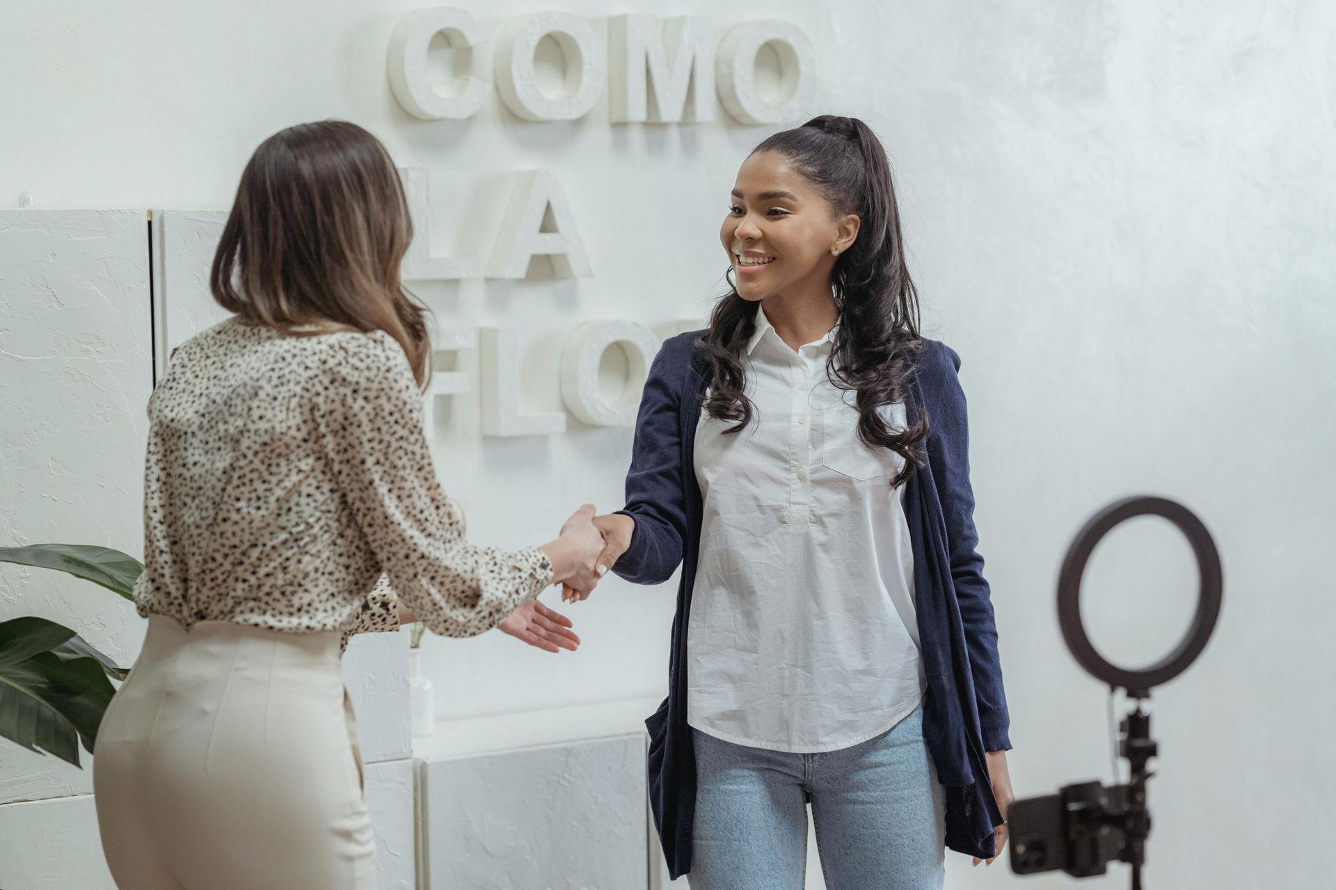 Two women shaking hands in a bright room with a ring light and wall text.