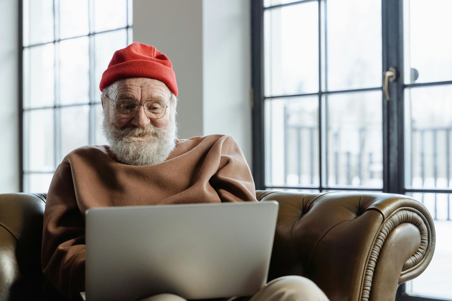 Man in red hat smiling at laptop, sitting on brown leather couch near a window.