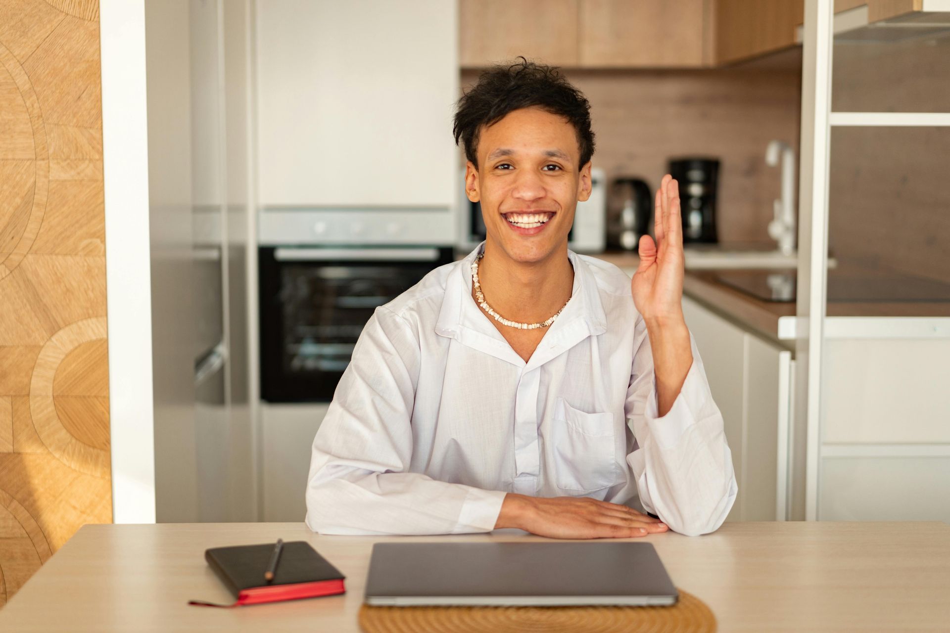 Person in a white shirt raises hand, smiling, seated at a desk with a laptop, notebook, and kitchen in the background.
