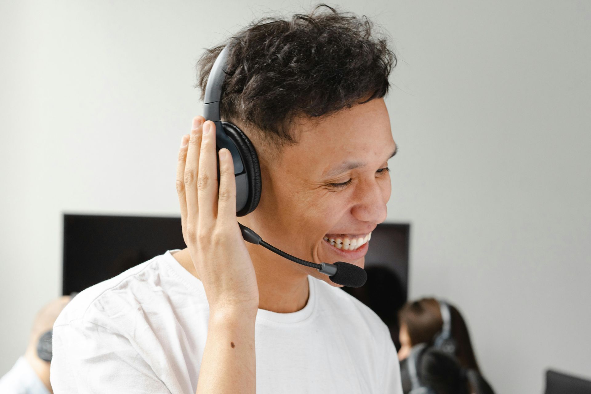 Man wearing a headset, smiling, touching ear. Light-colored room, possibly an office, with others in the background.