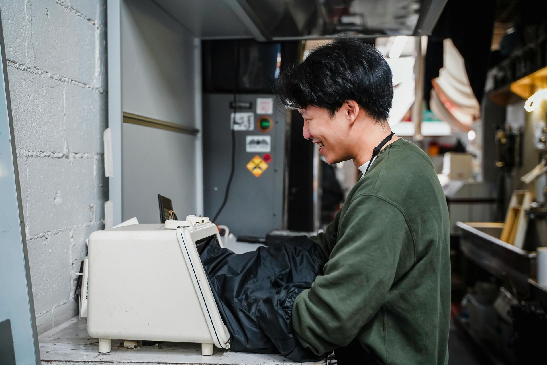 Man in green sweater, smiling, operates a machine inside a workshop.