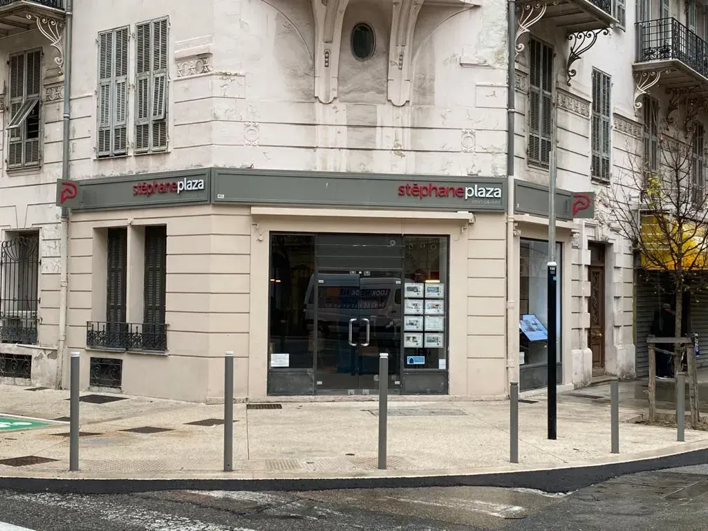 Façade d'un immeuble d'angle avec une enseigne grise pour Stéphane Plaza Immobilier, avec portes vitrées et vitrines.