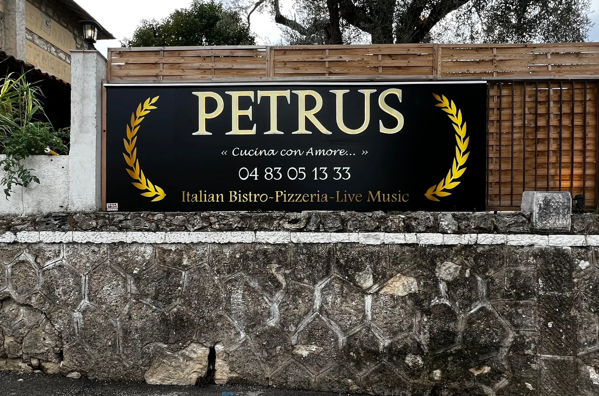 Une enseigne noire pour le restaurant italien Petrus, avec des lettres dorées et des couronnes de laurier, fixée sur un mur de pierre.