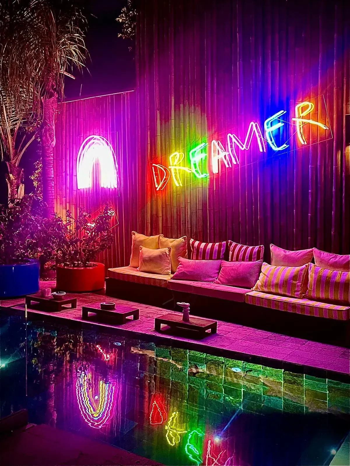 Un salon extérieur lumineux avec une enseigne lumineuse « DREAMER » et un arc-en-ciel se reflétant dans une piscine la nuit.