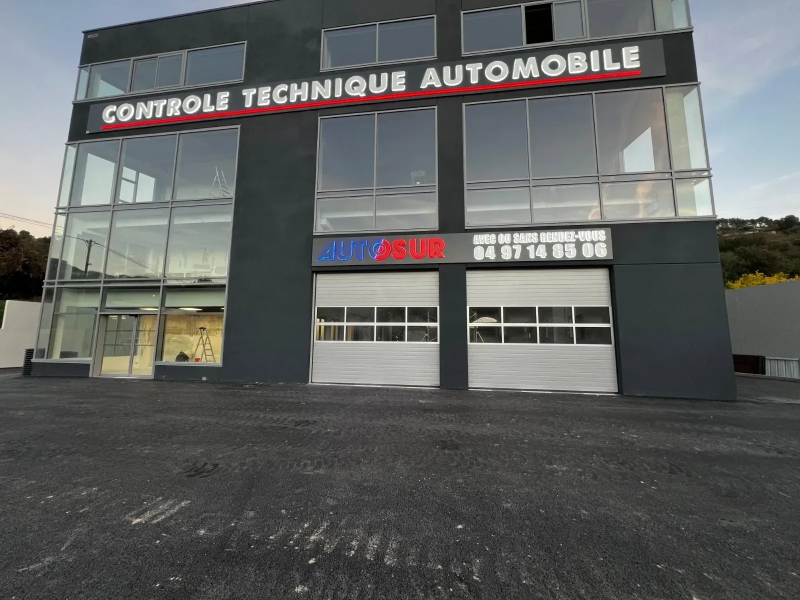 Un bâtiment commercial moderne, gris foncé, avec de grandes fenêtres et des portes de garage blanches à enroulement, portant l'inscription « Contrôle Technique ».