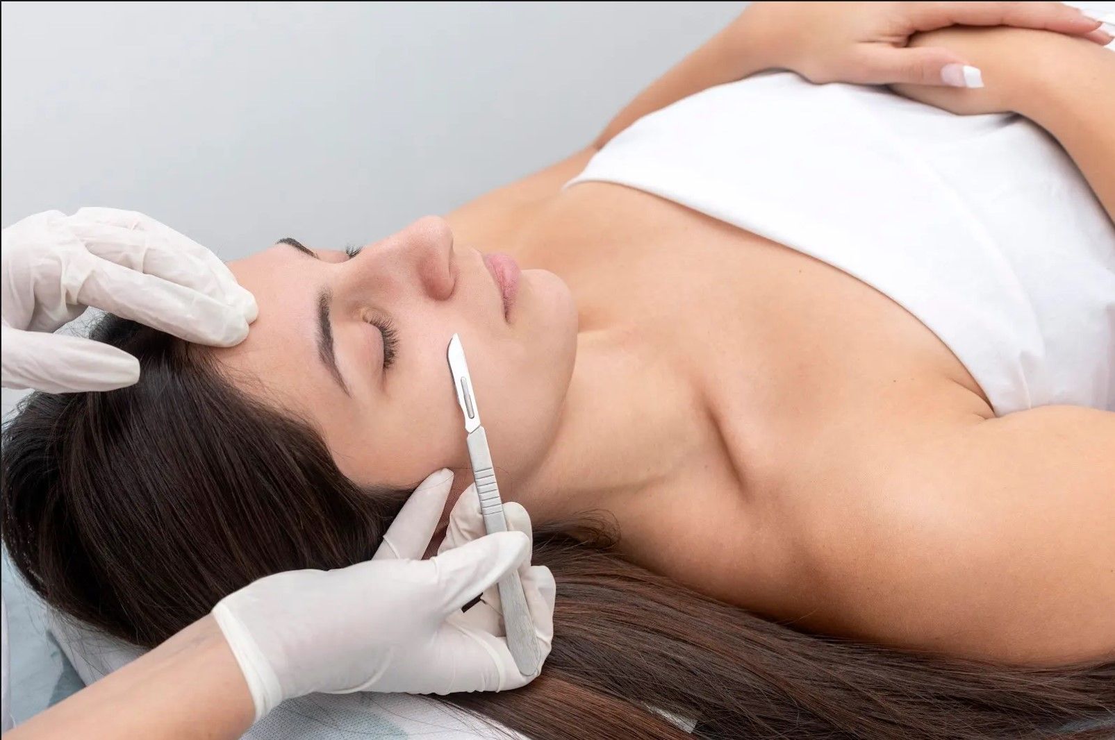 Woman undergoing a cosmetic procedure with a scalpel on her face while laying on a medical table.