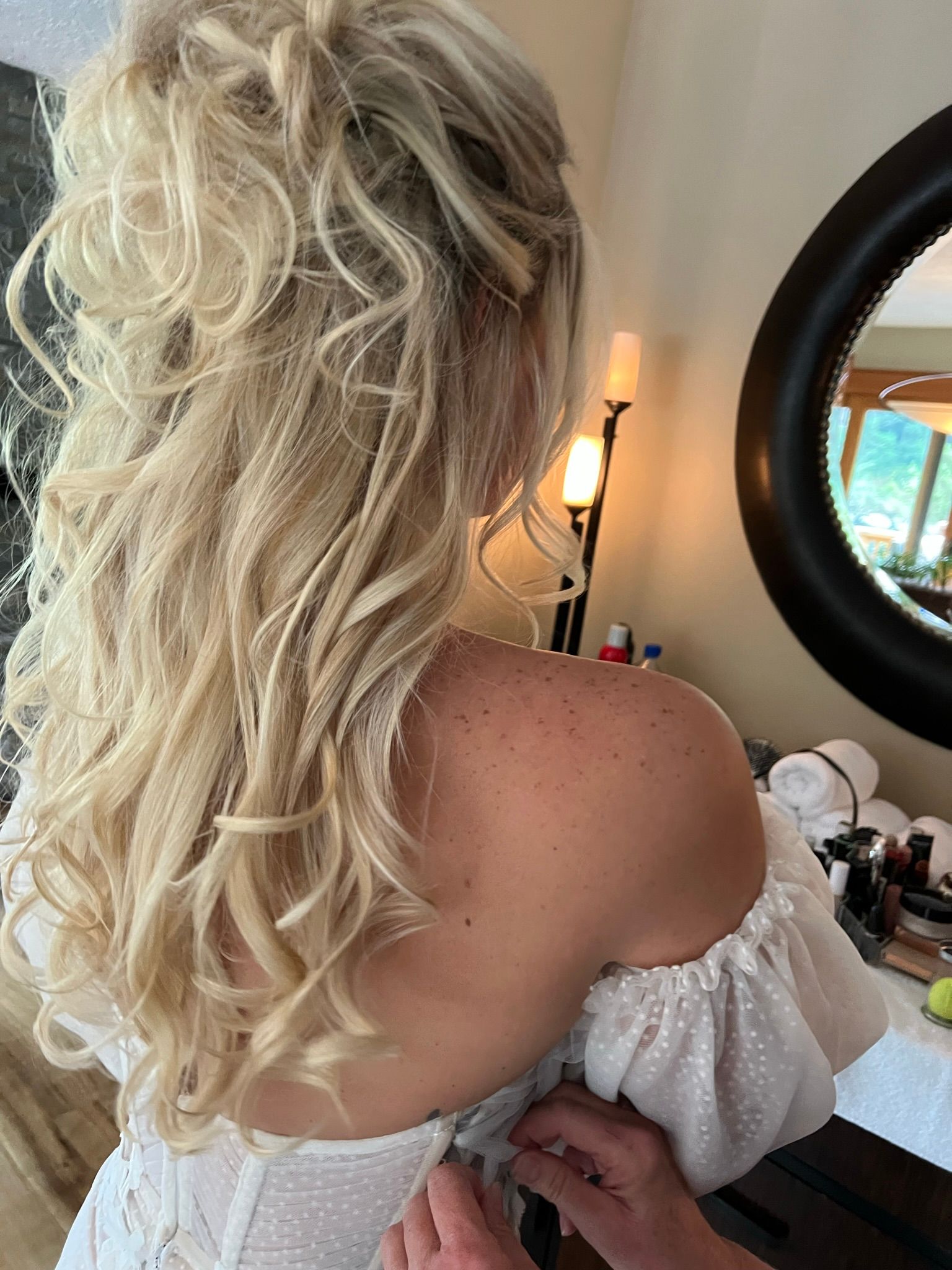 A woman in a white dress is getting her hair done in front of a mirror.