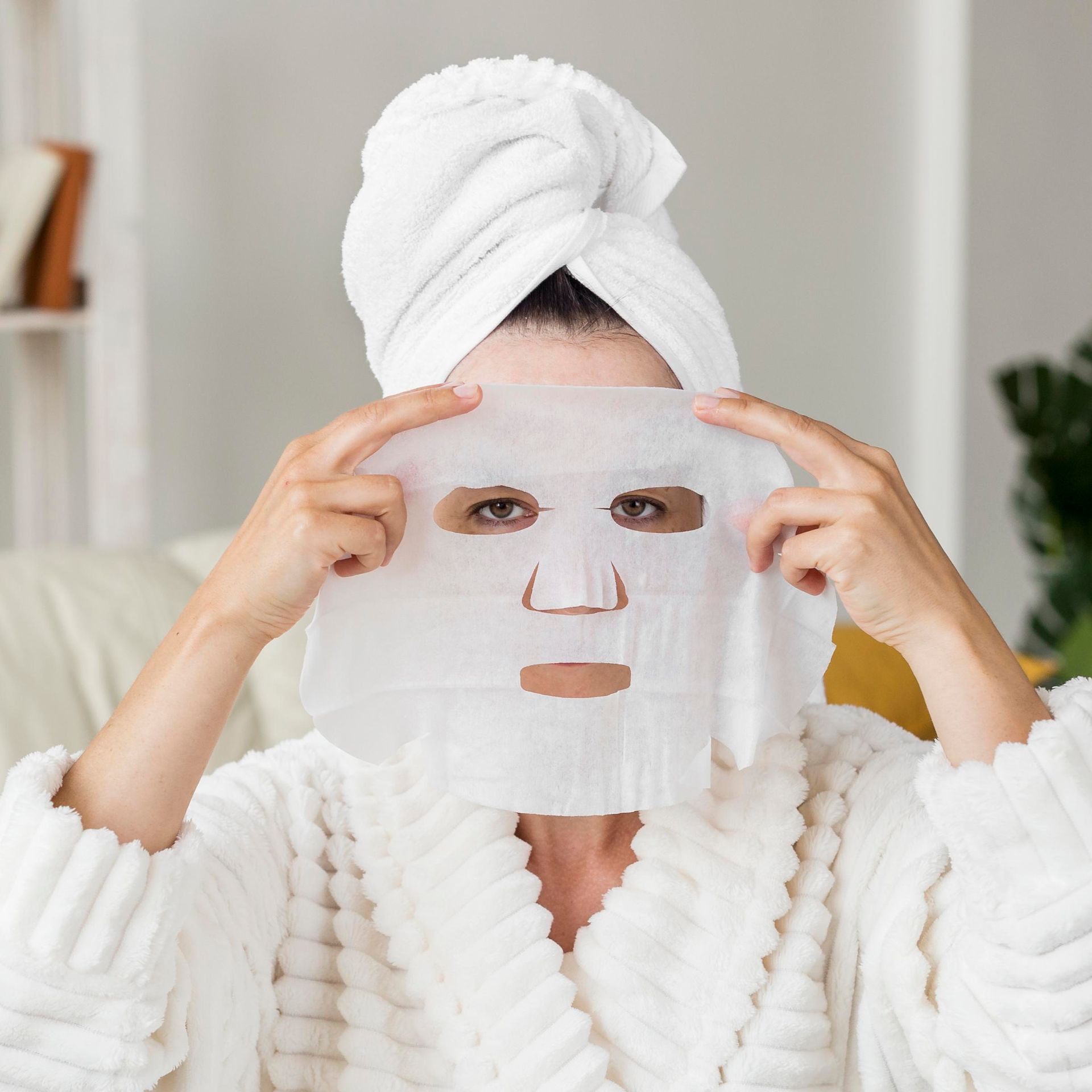 A woman with a towel wrapped around her head is covering her face with a sheet mask.