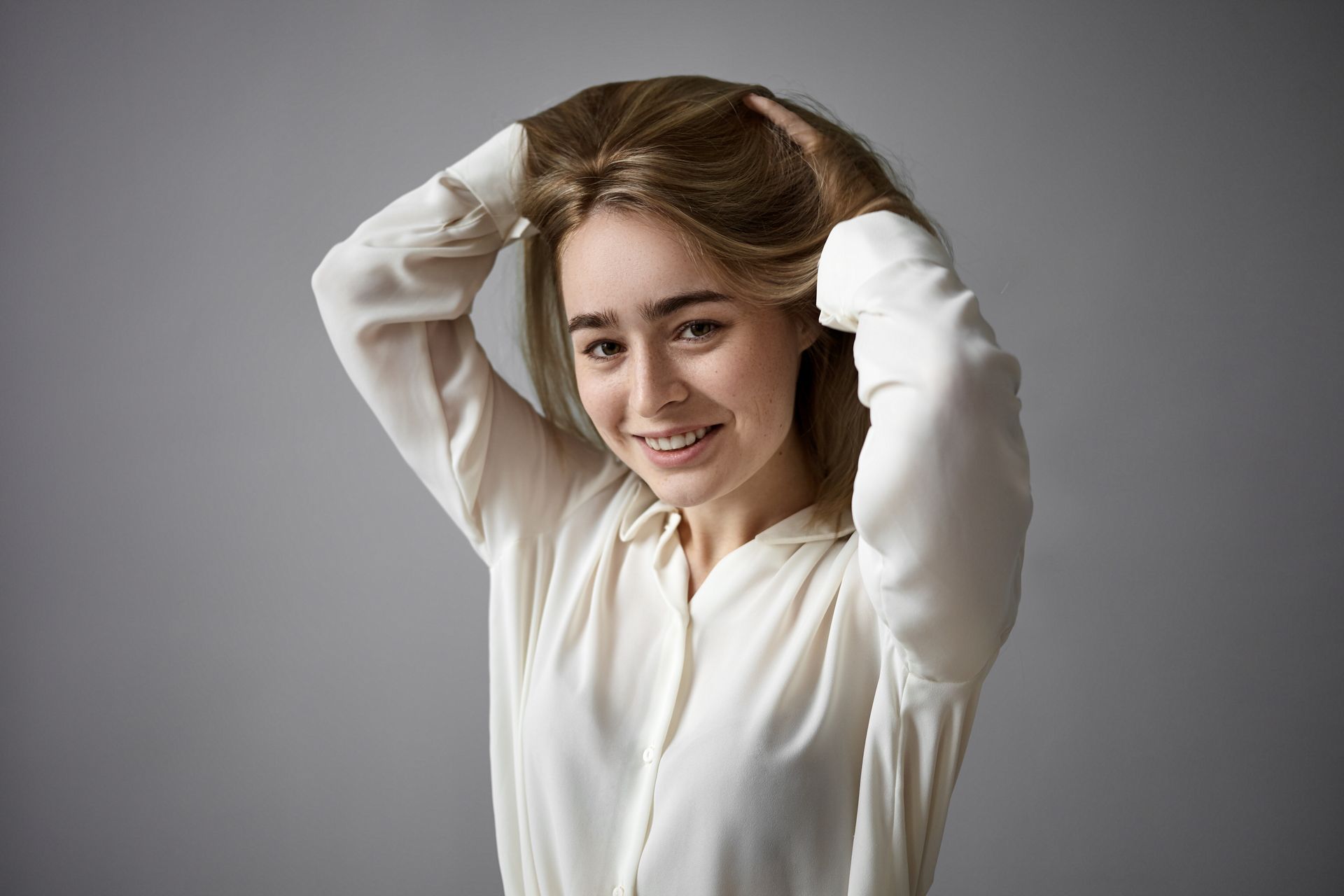 A woman in a white shirt is smiling and holding her hair.