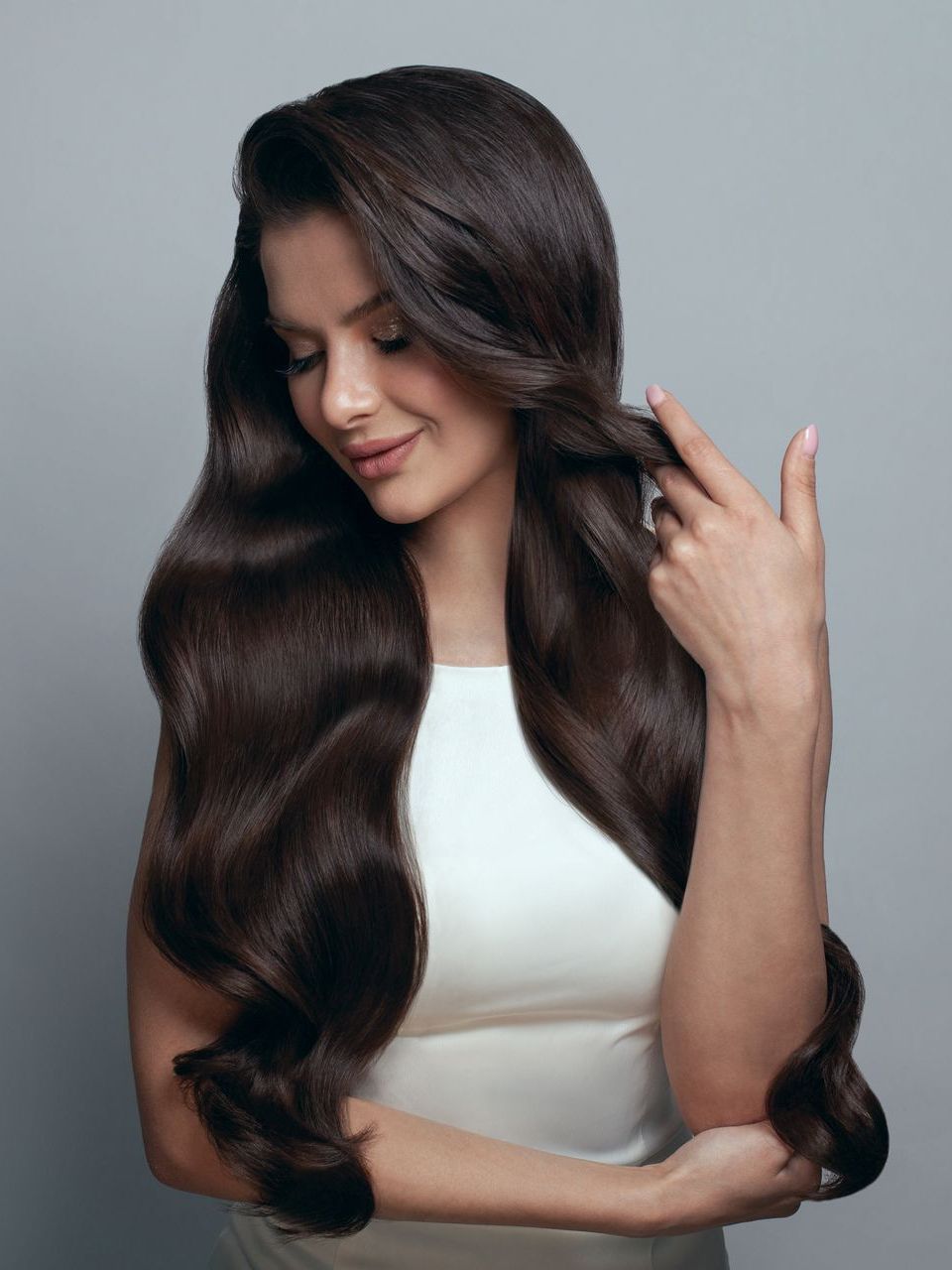 A woman with long brown hair is wearing a white dress and touching her hair.