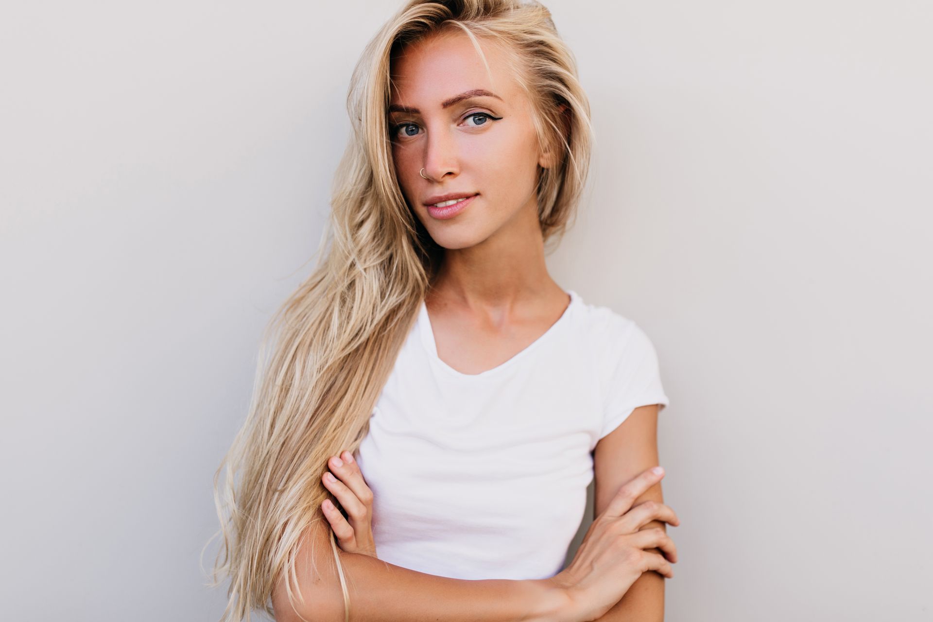 A woman with long blonde hair is wearing a white t-shirt and standing with her arms crossed.