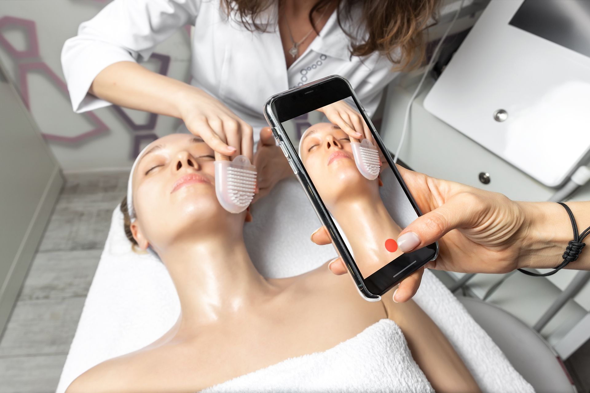 A woman is getting a facial treatment at a spa and a person is taking a picture of her face.