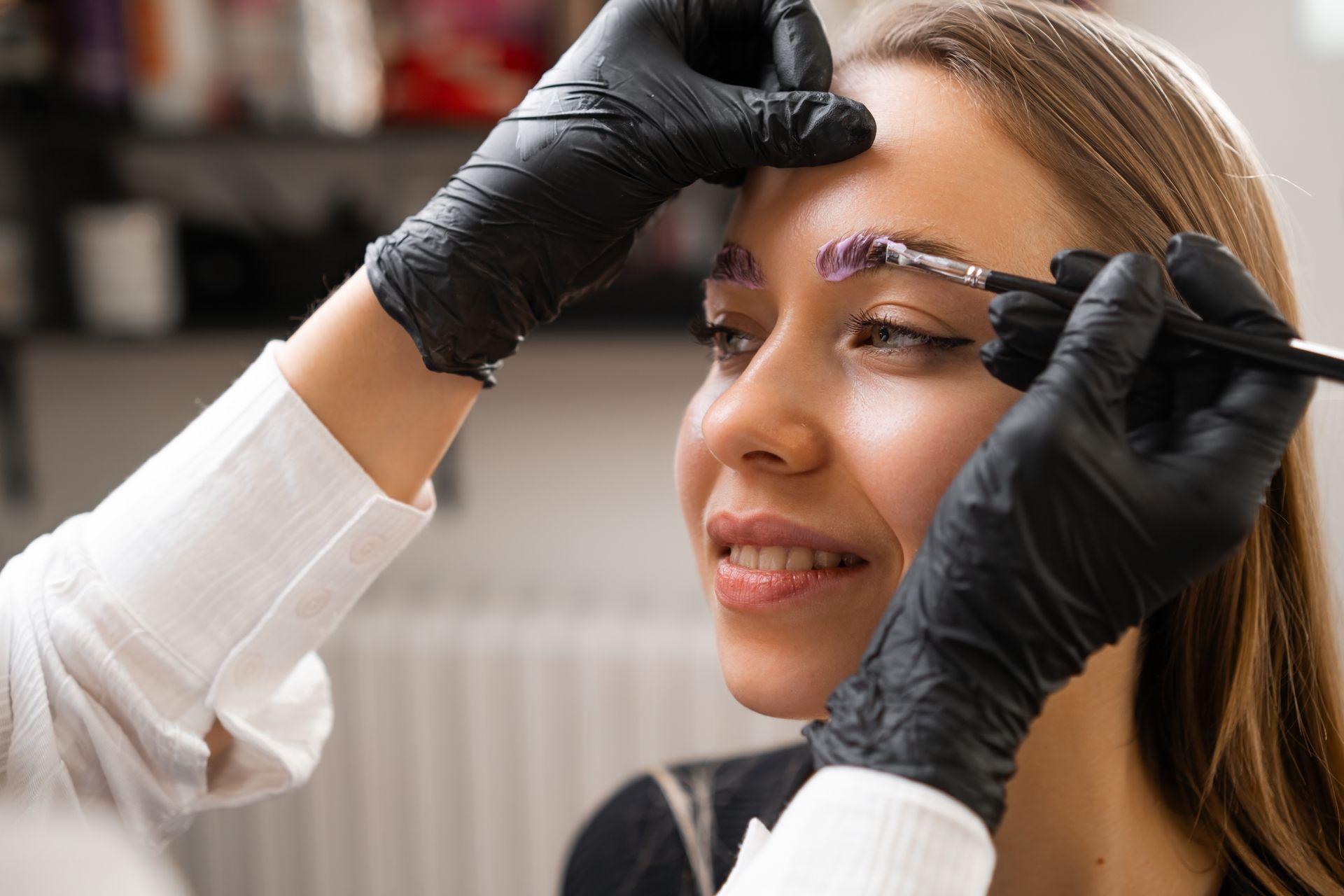 A beauty professional wearing black gloves uses a small brush to apply product to a client’s eyebrow.