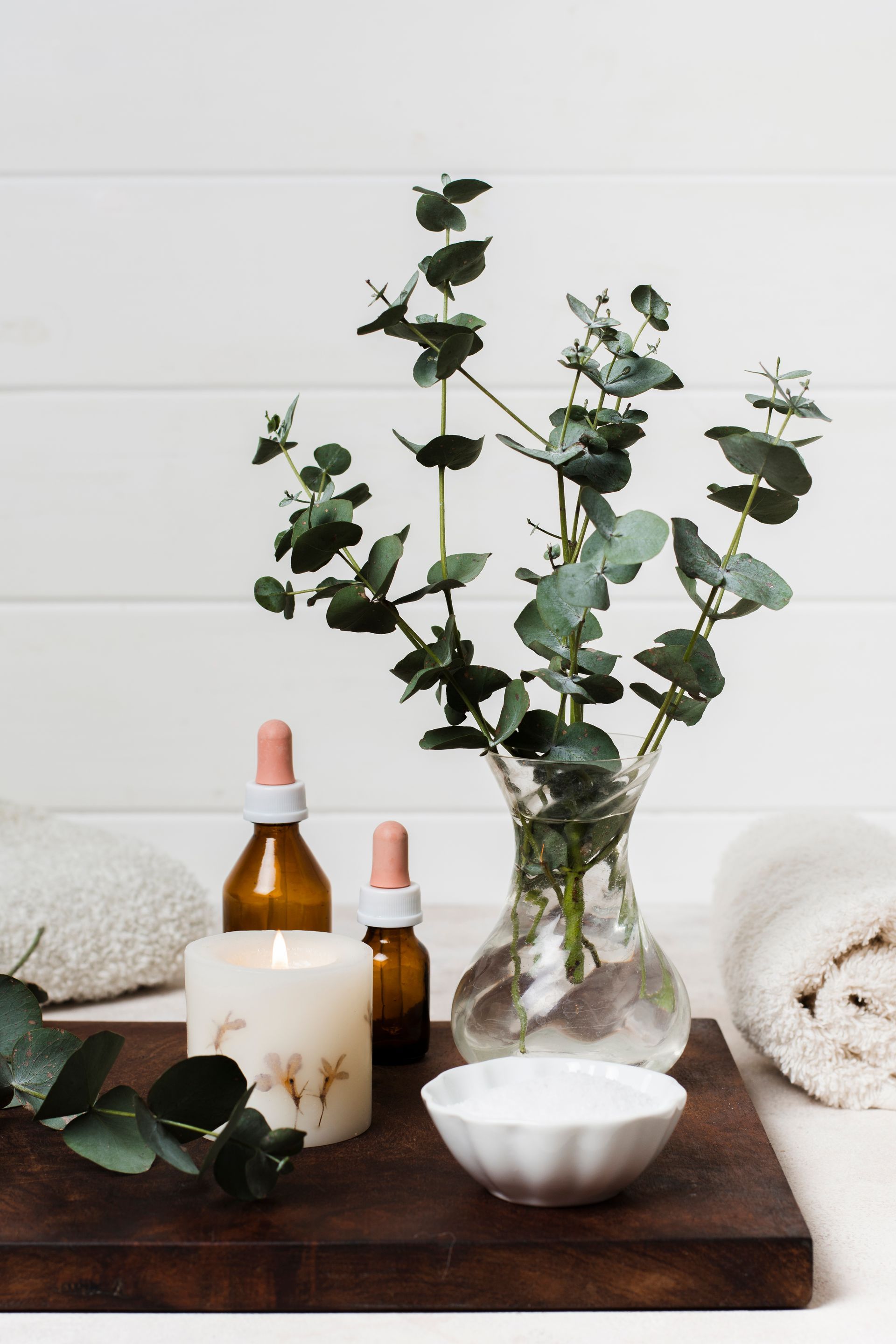 A wooden tray with a vase of eucalyptus , a candle , and bottles of essential oils.